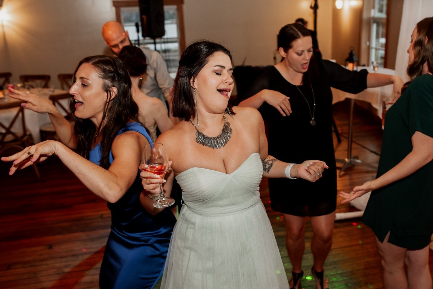 Guests dance joyfully at an indoor party while one person gracefully holds a wine glass, capturing a moment of elegance and celebration, resembling a wedding scene at The Loft at the Red Building.