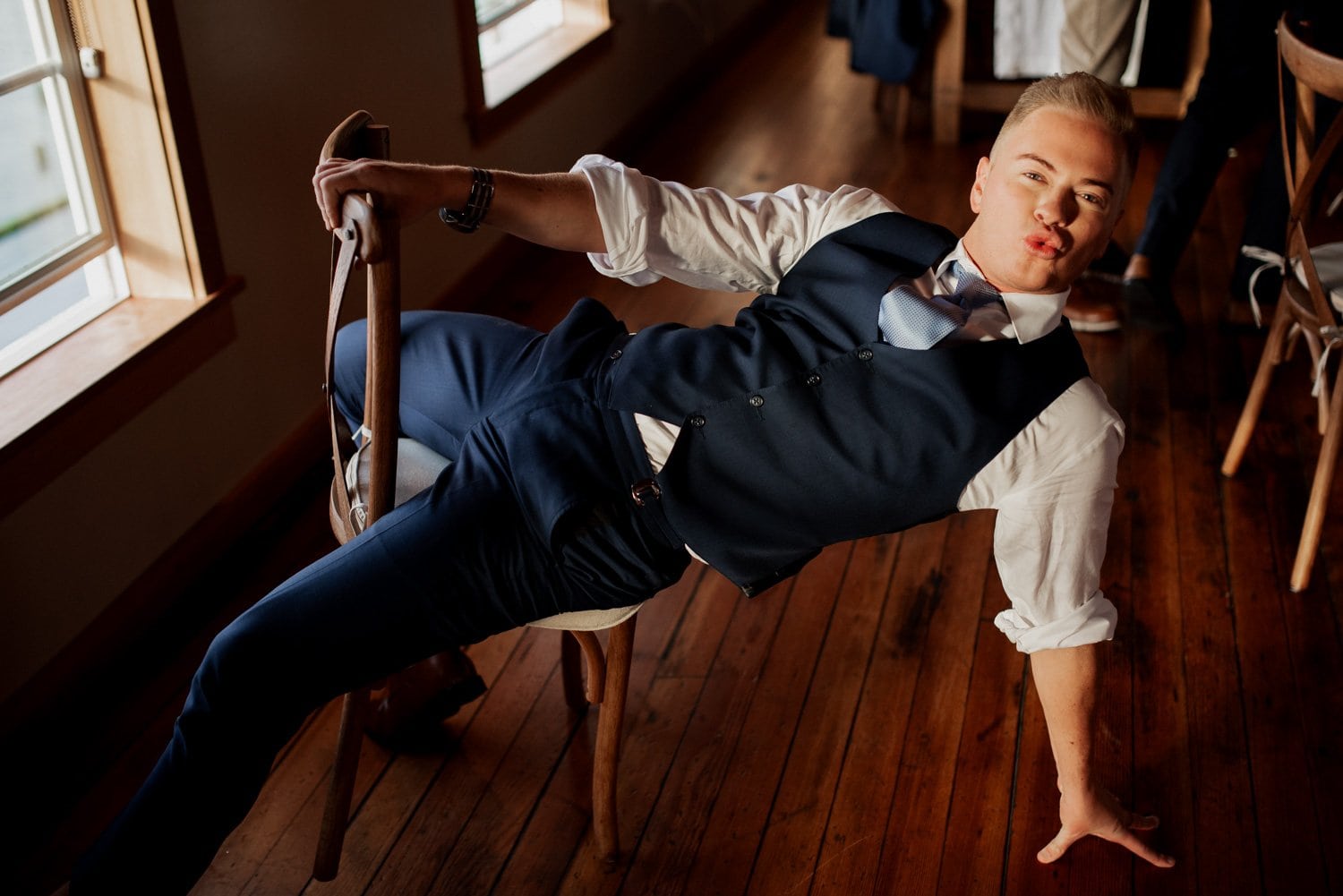 A person in a suit lounges casually on a chair, touching the wooden floor with one hand in a warmly lit room.