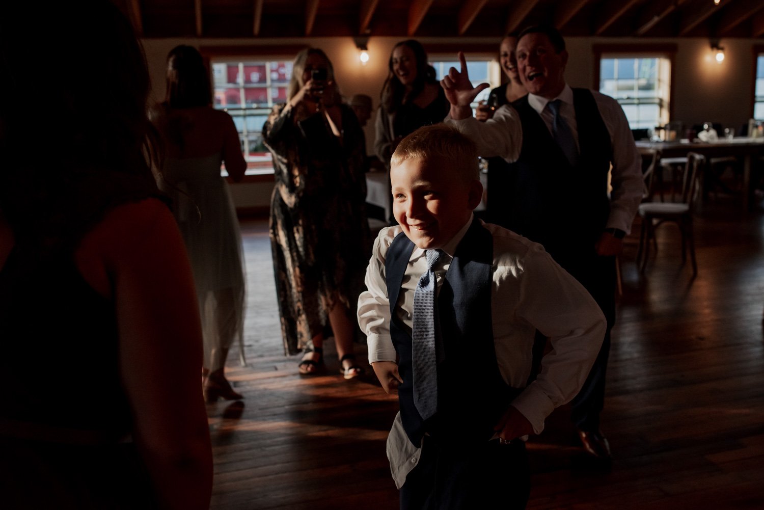 A young boy in formal attire dances energetically at a wedding, with adults around him cheering and taking photos under the bright lights of The Loft at the Red Building.