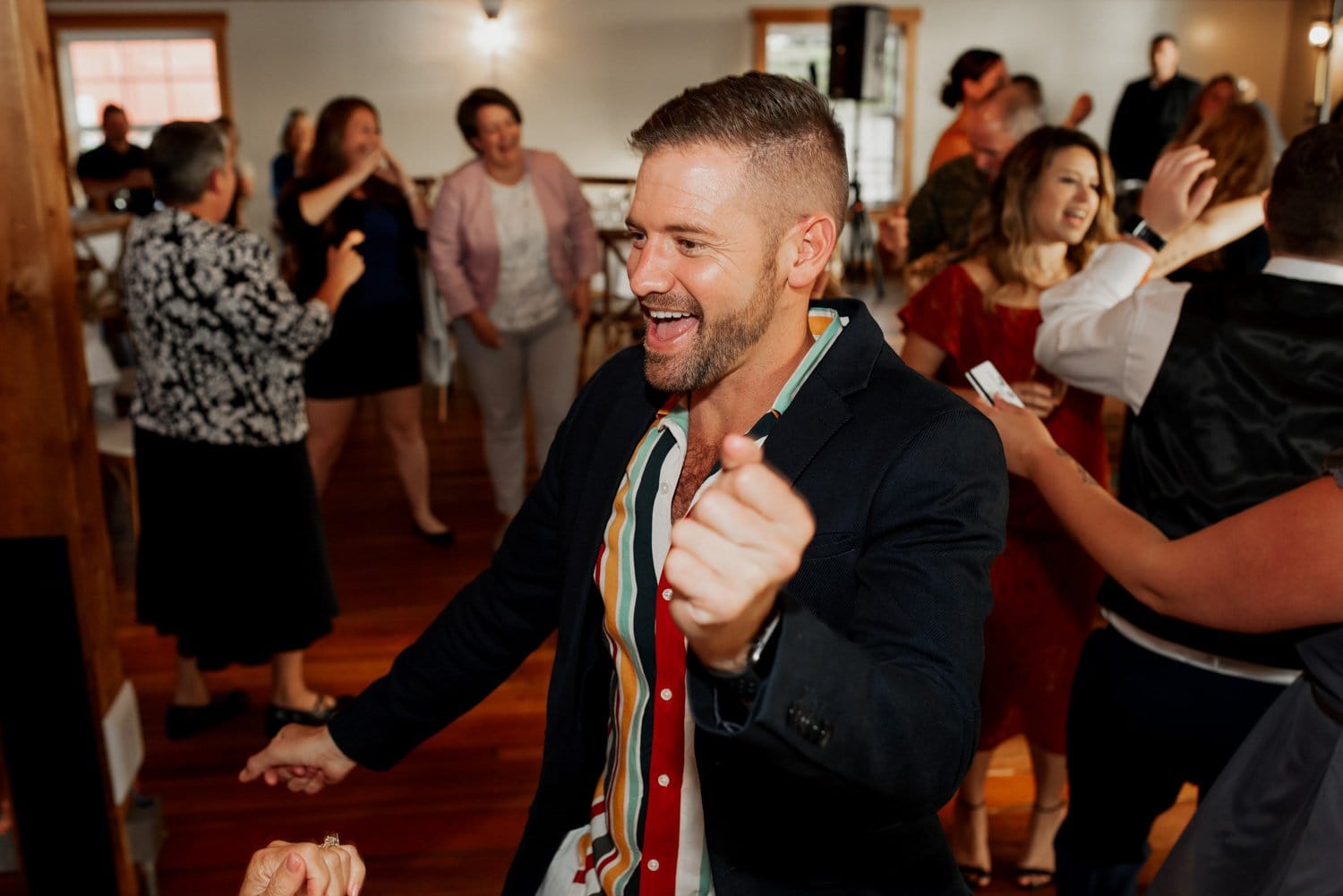 A man in a suit joyfully dances at an indoor wedding at The Loft at the Red Building, with other guests celebrating around him.