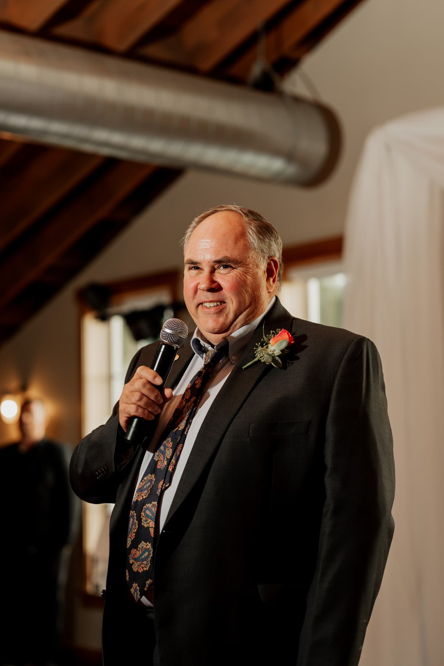 A man in a suit with a floral tie and boutonniere holds a microphone, smiling in a warmly lit room. This wedding photo captures an intimate moment from the ceremony at The Loft at the Red Building.