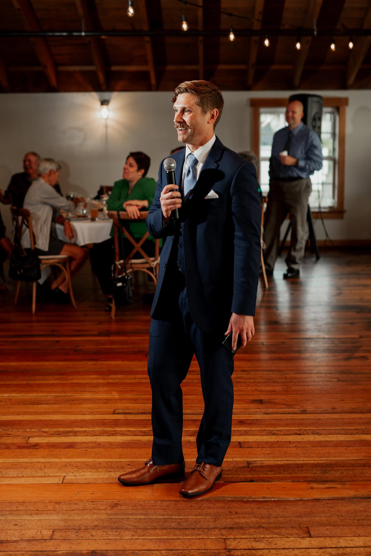 A man in a suit speaks into a microphone indoors as guests seated at tables watch with warmth. The scene is part of wedding photos taken at The Loft at the Red Building, showcasing both elegance and emotion.