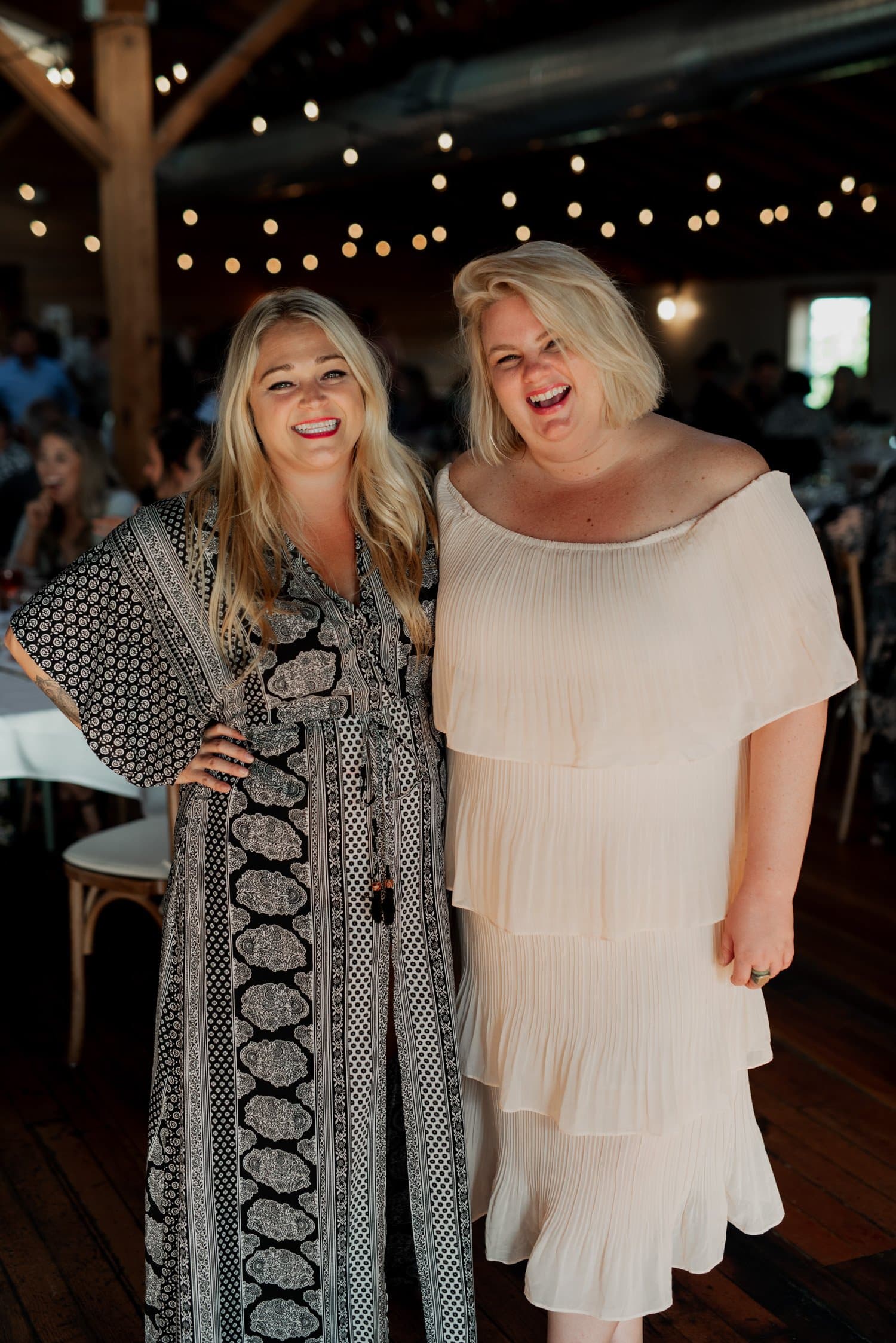 Two women smiling and standing together indoors with string lights twinkling in the background at a wedding at The Loft at the Red Building.