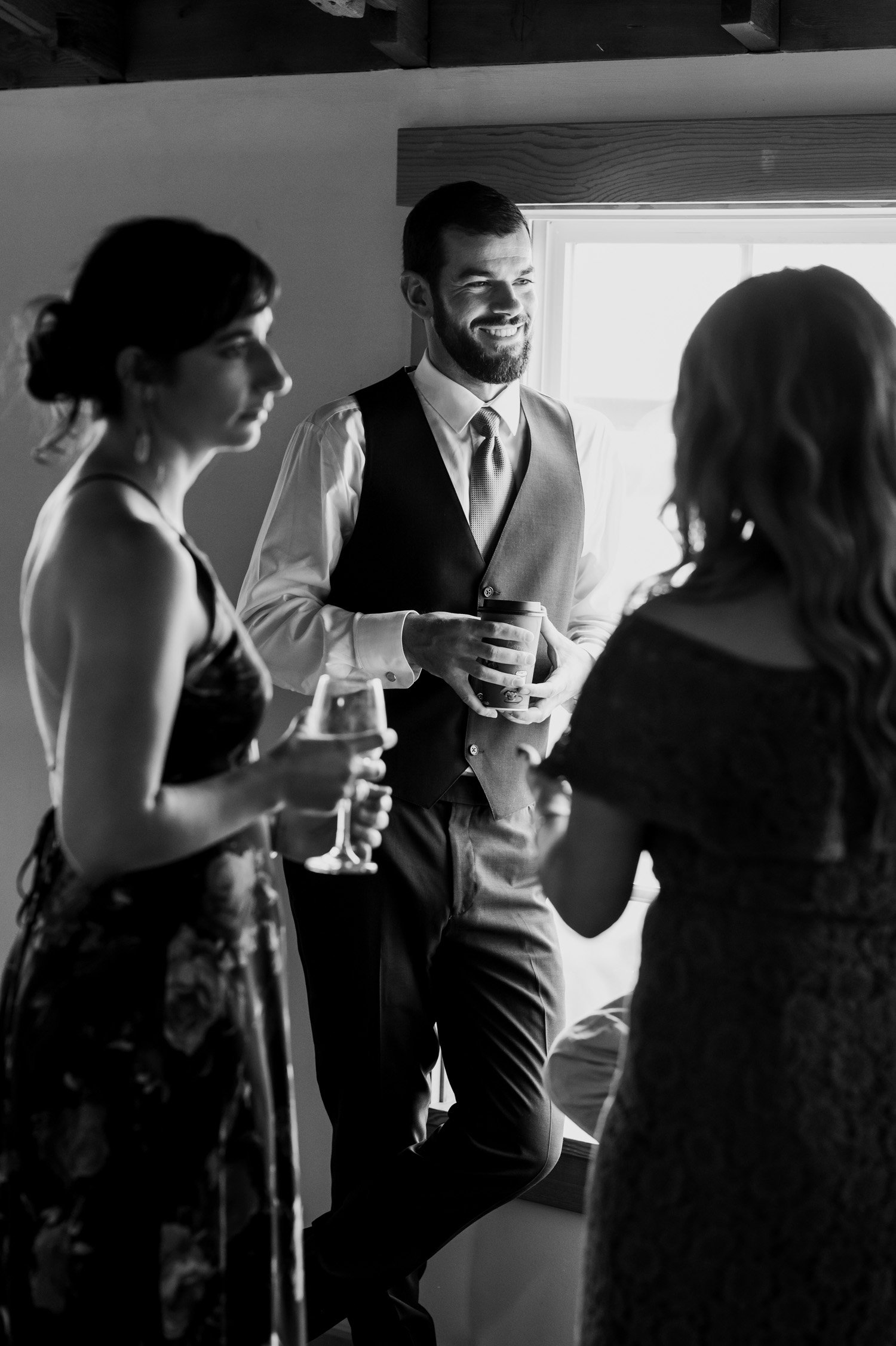 Three people in formal attire stand by a bright window, holding drinks and engaging in conversation at The Loft at the Red Building.