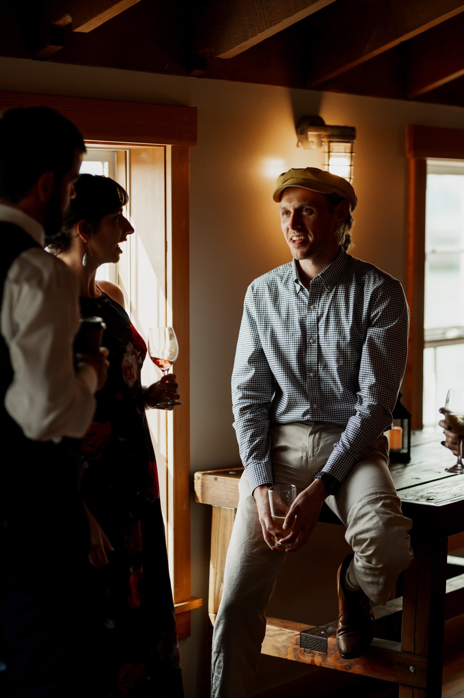 A person wearing a cap sits casually on a table, conversing with two others who are holding drinks in a warmly lit room during wedding photos at The Loft at the Red Building.