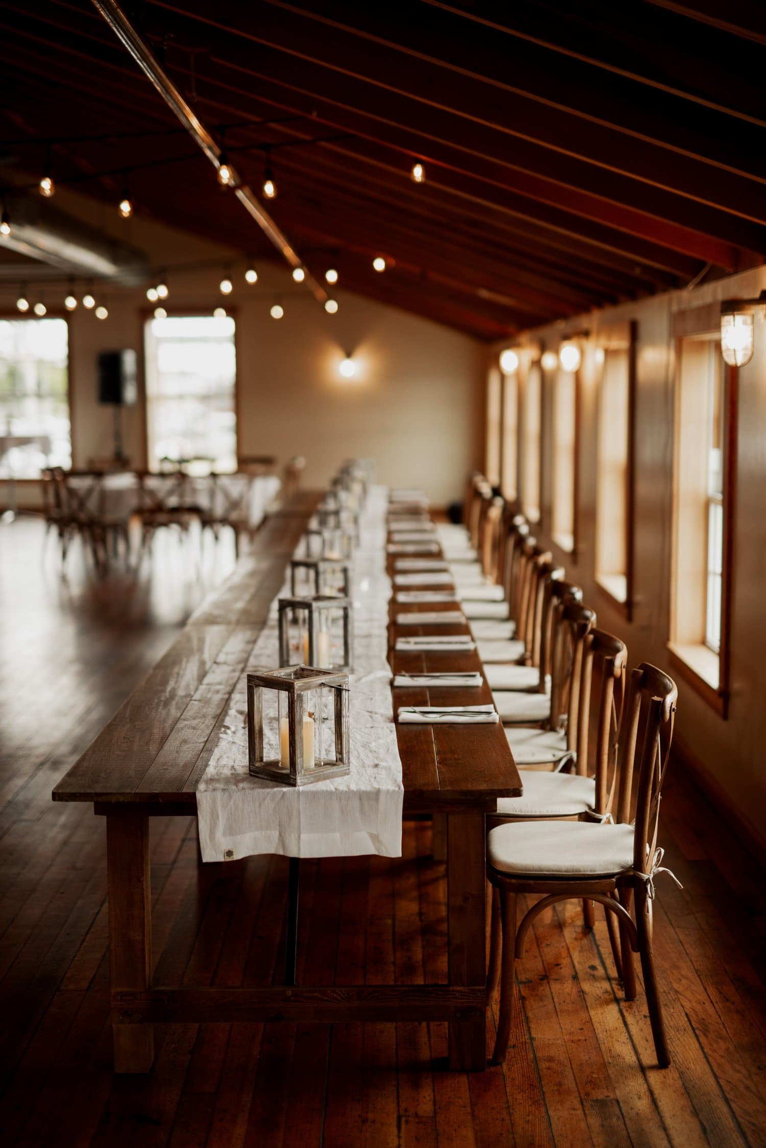 A long wooden table with chairs, lanterns, and a white runner in The Loft at the Red Buildings rustic room. Warm lighting and a charming wooden floor enhance the setting.