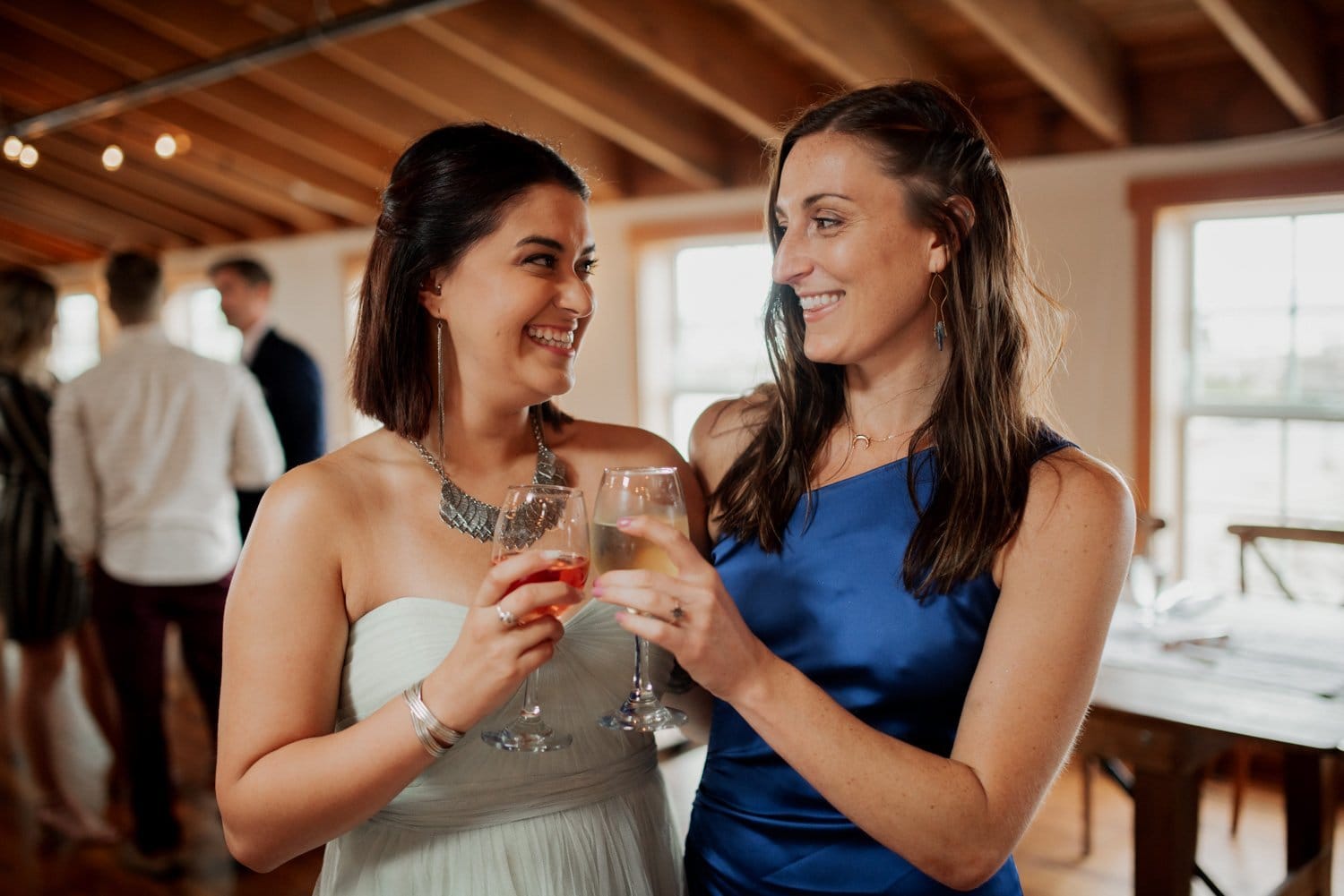 Two women in elegant dresses smile and toast with wine glasses in a warmly lit room with wooden ceilings.