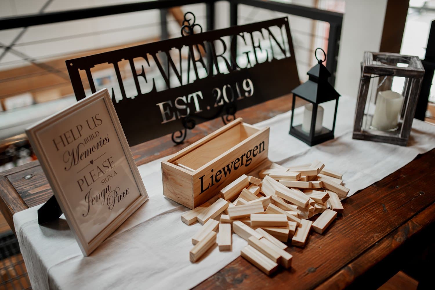 A table display featuring wooden blocks, a candle lantern, and a framed message about building memories. Next to it is a sign that reads Liewergen Est. 2019, creating an inviting and nostalgic decor piece.