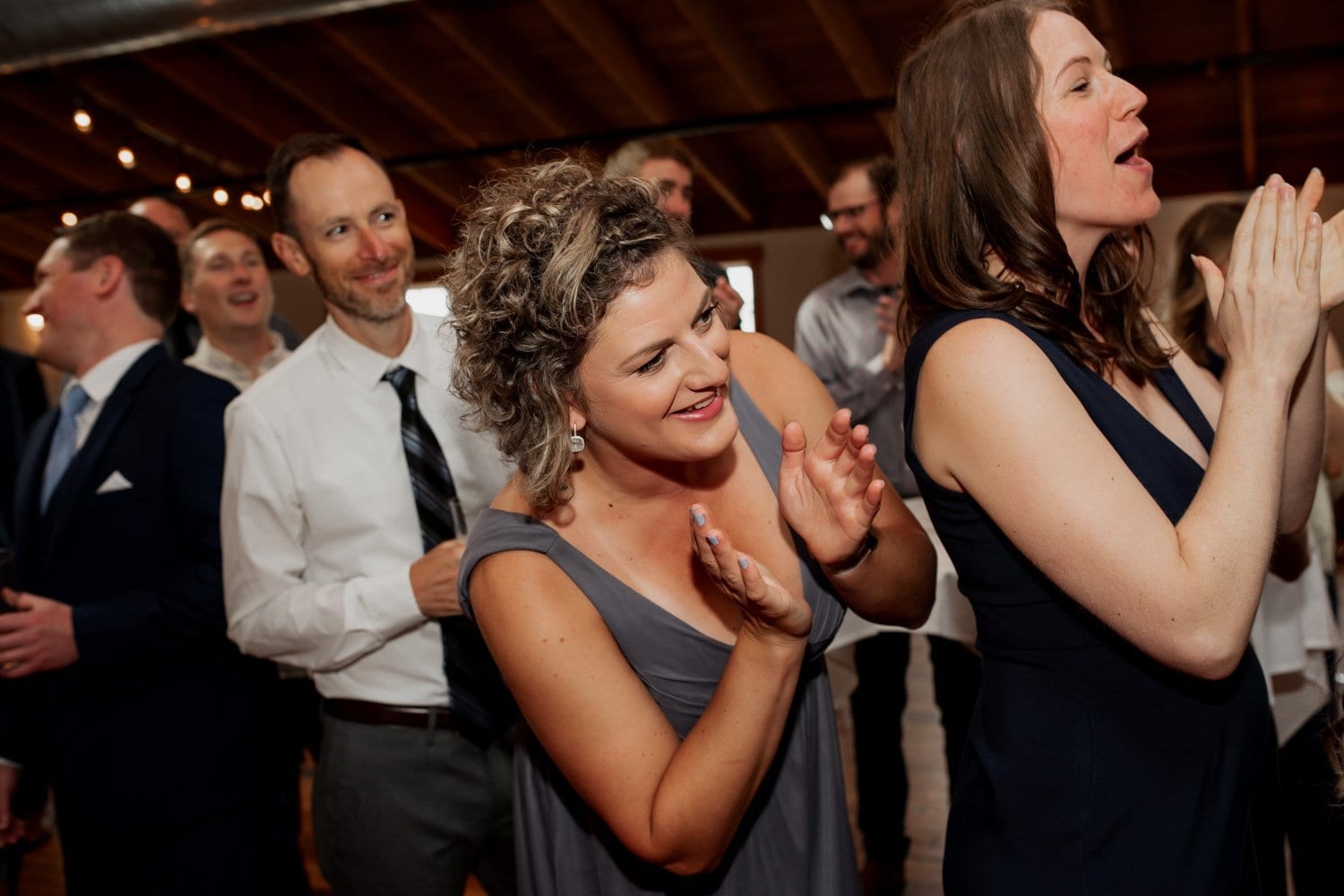 A group of people clapping and smiling at a wedding held at The Loft at the Red Building, with wooden ceilings and string lights enhancing the atmosphere.