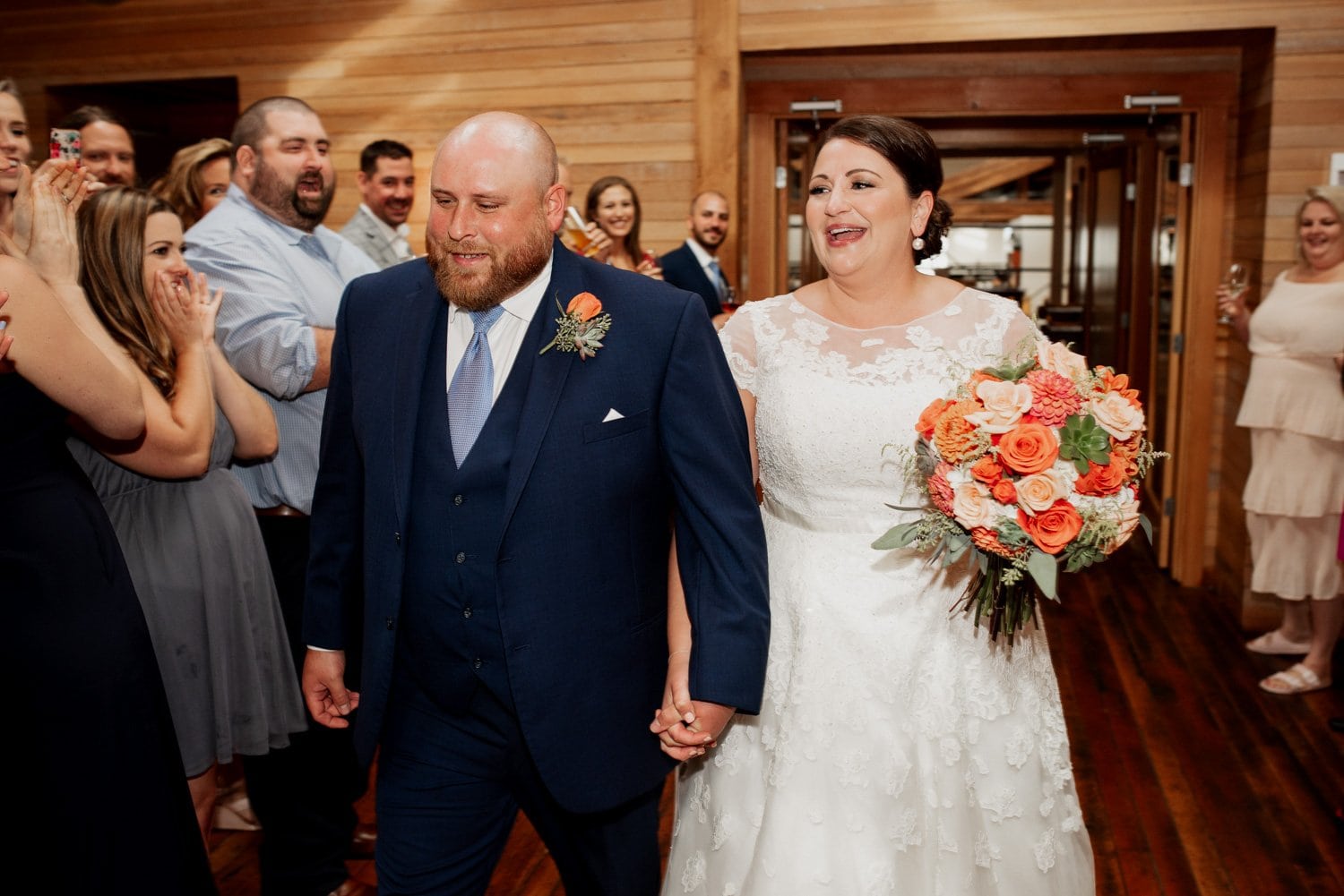 Bride and groom holding hands and smiling; friends cheer in a wooden hall during their wedding at The Loft at the Red Building.