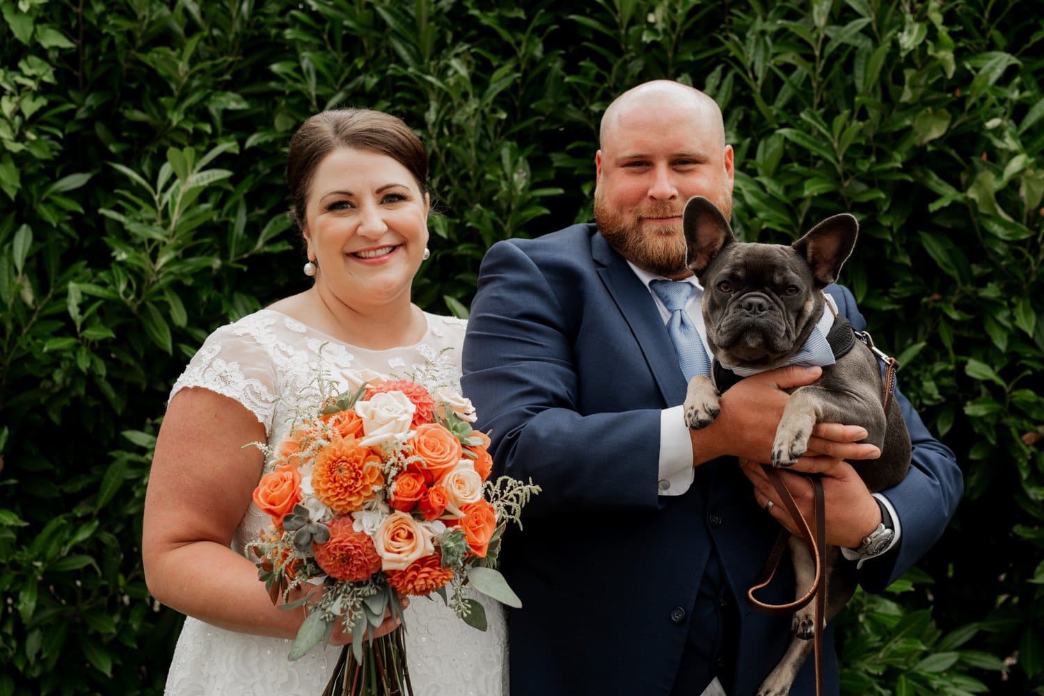 A smiling couple in wedding attire holds a dog against a backdrop of lush green foliage at Astorias Maritime Memorial Park. The bride holds a bouquet of orange flowers, suggesting their recent wedding celebration.