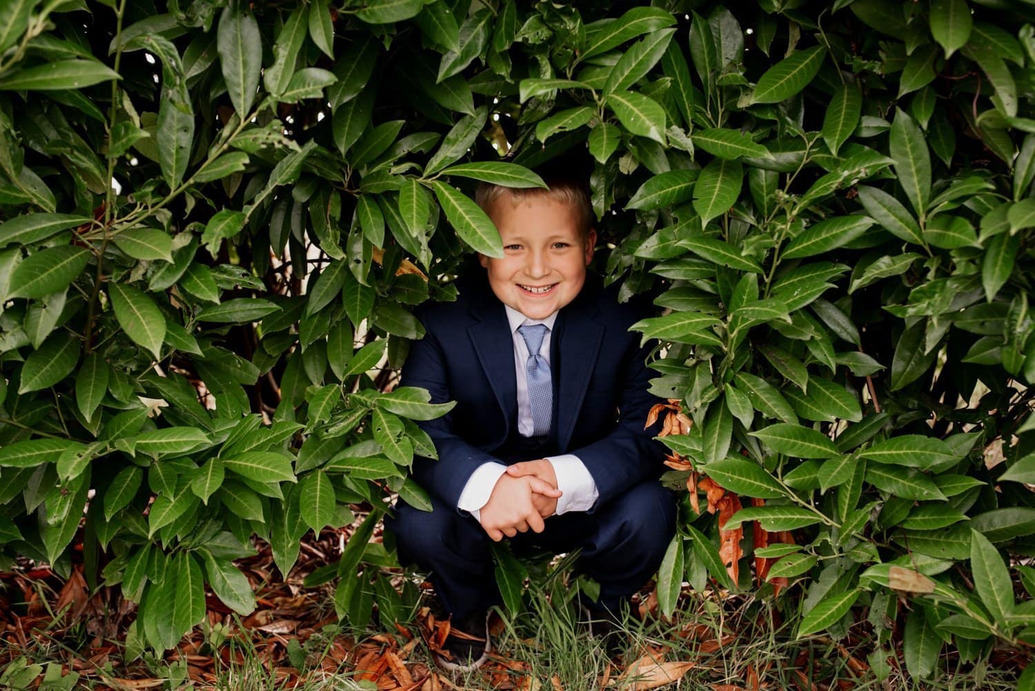 A child in a suit crouches and smiles within an opening in a leafy bush at Astorias Maritime Memorial Park, just before a wedding ceremony.