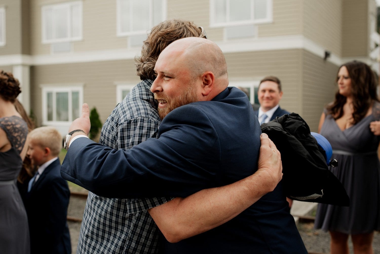 Two men hugging at an outdoor wedding ceremony with guests in formal attire at Astorias Maritime Memorial Park.