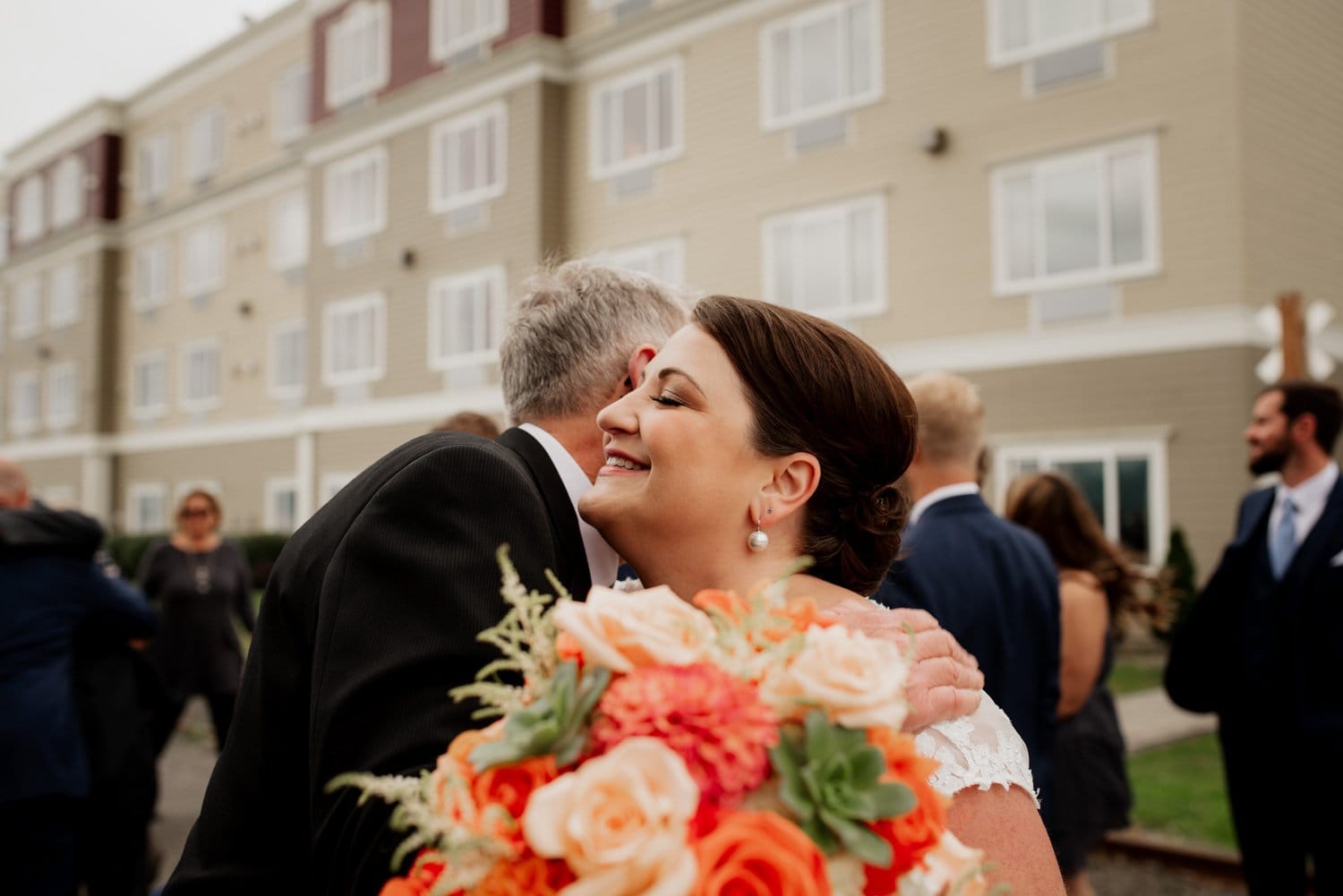 A bride holding her bouquet embraces a man affectionately in front of a building at Astorias Maritime Memorial Park, with guests congregating in the background.