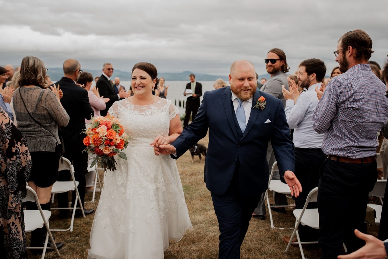 A bride and groom walk hand in hand down an outdoor aisle at Astorias Maritime Memorial Park, smiling as they are surrounded by clapping guests.