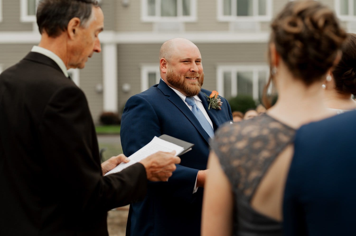A groom in a blue suit smiles during a wedding ceremony at Astorias Maritime Memorial Park, with people standing nearby.