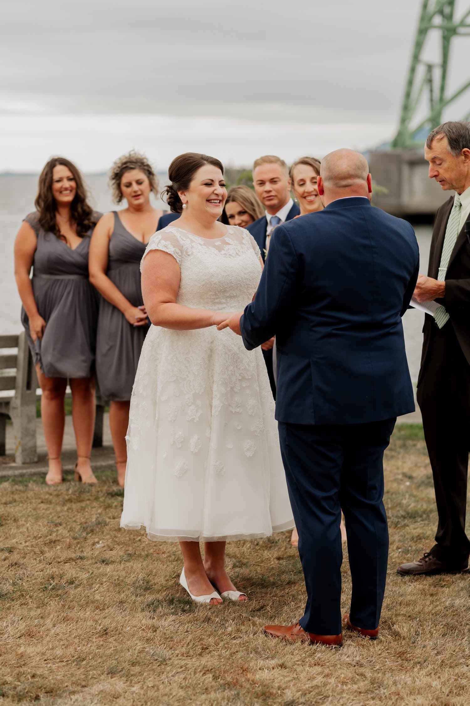 A bride and groom hold hands during their wedding ceremony at Maritime Memorial Park in Astoria, surrounded by bridesmaids with a bridge in the background.