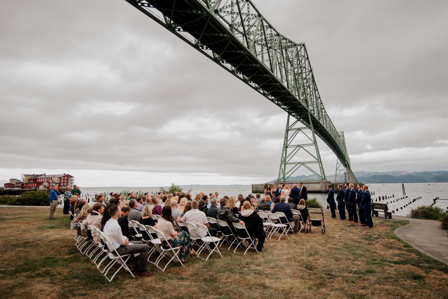 An outdoor wedding ceremony is held at Maritime Memorial Park in Astoria, beneath a large bridge. Guests are seated in rows under a cloudy sky, with calm water visible in the background.