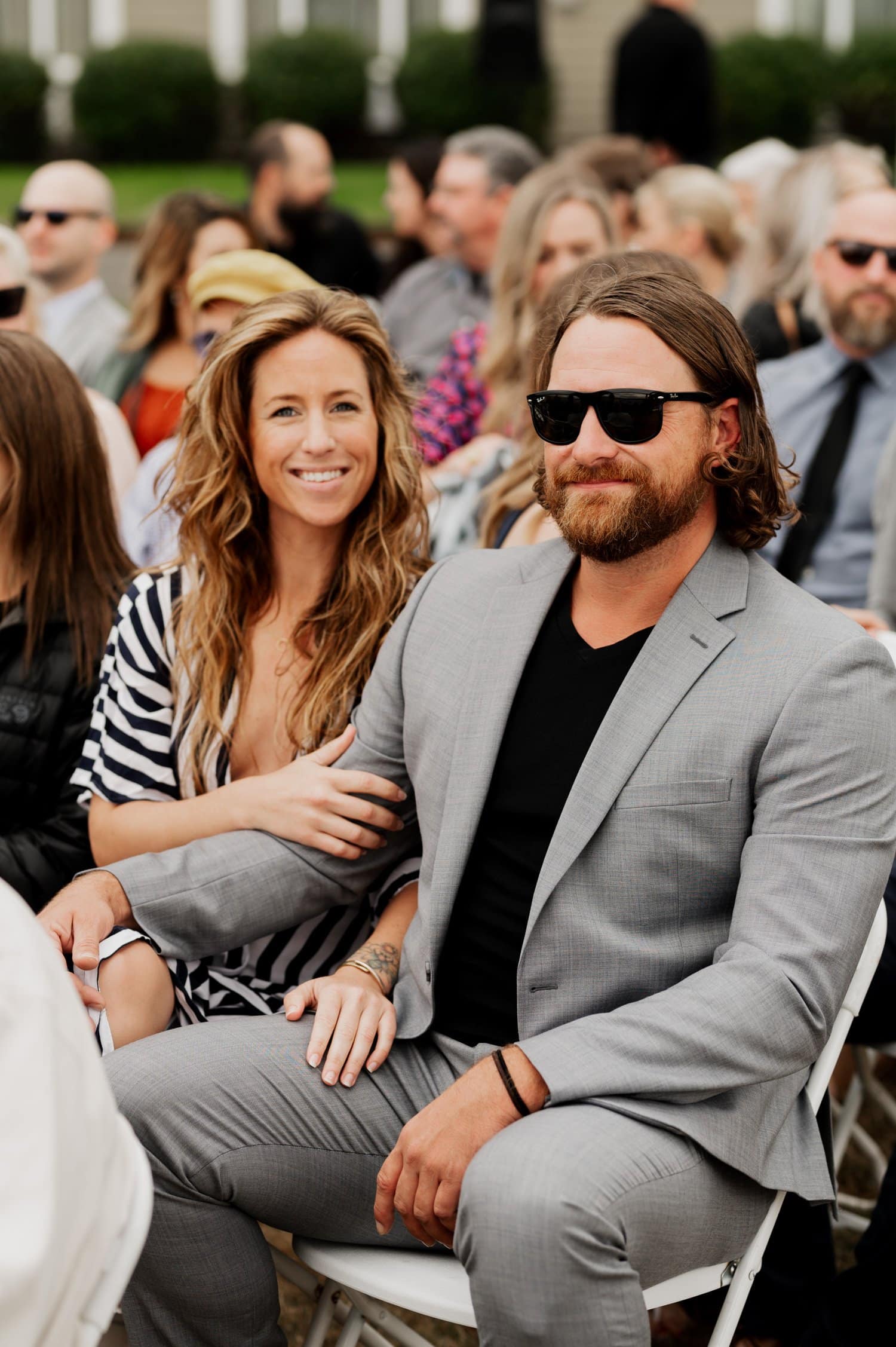A smiling couple sits together at an outdoor event, possibly a wedding ceremony at Astorias Maritime Memorial Park. The man is dressed in a gray suit, and the woman wears a striped outfit. They appear to be looking forward to attending the reception at The Loft at the Red Building.