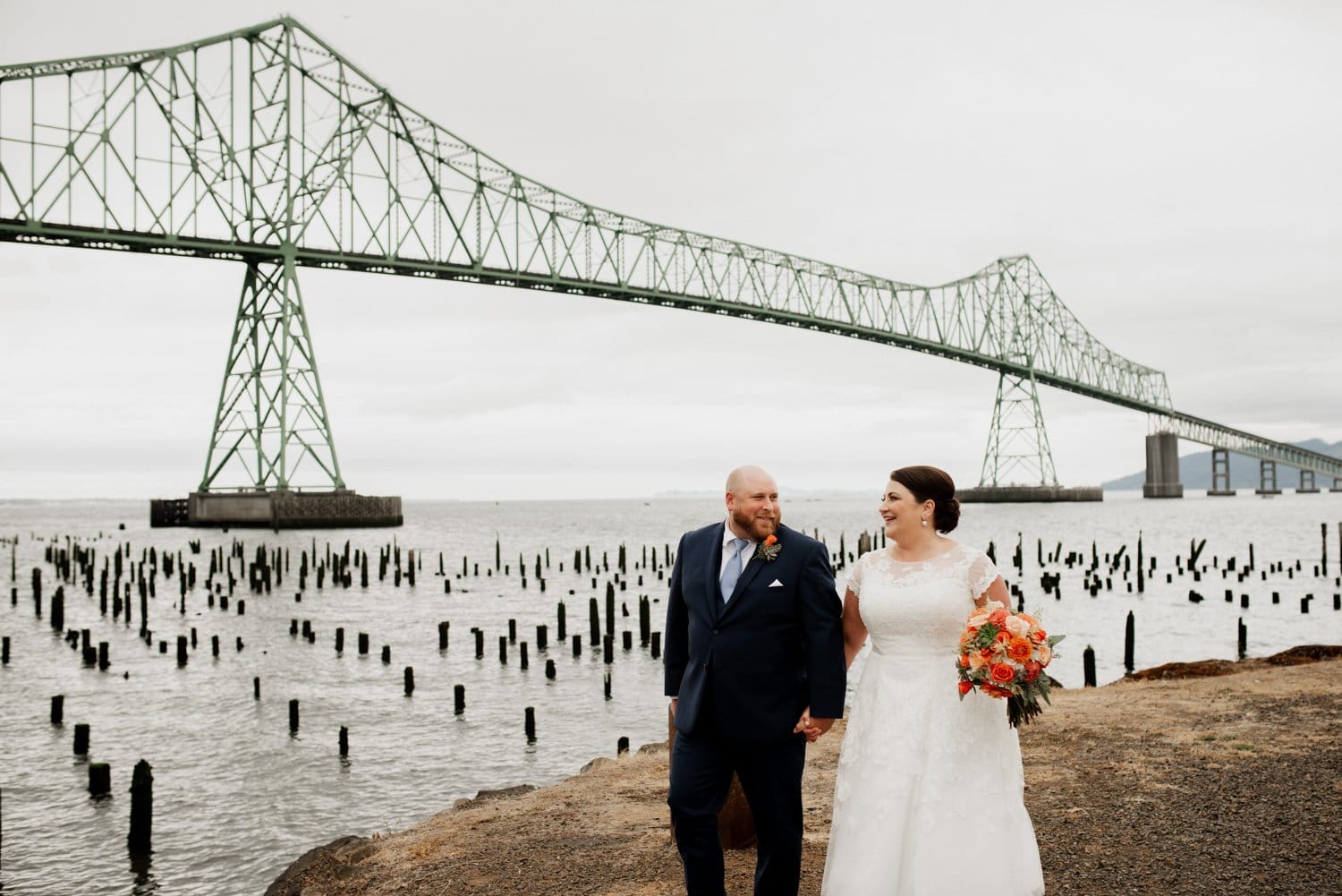 A bride and groom hold hands near a large bridge with wooden pilings in the water, capturing perfect wedding photos for their ceremony at The Loft at the Red Building.