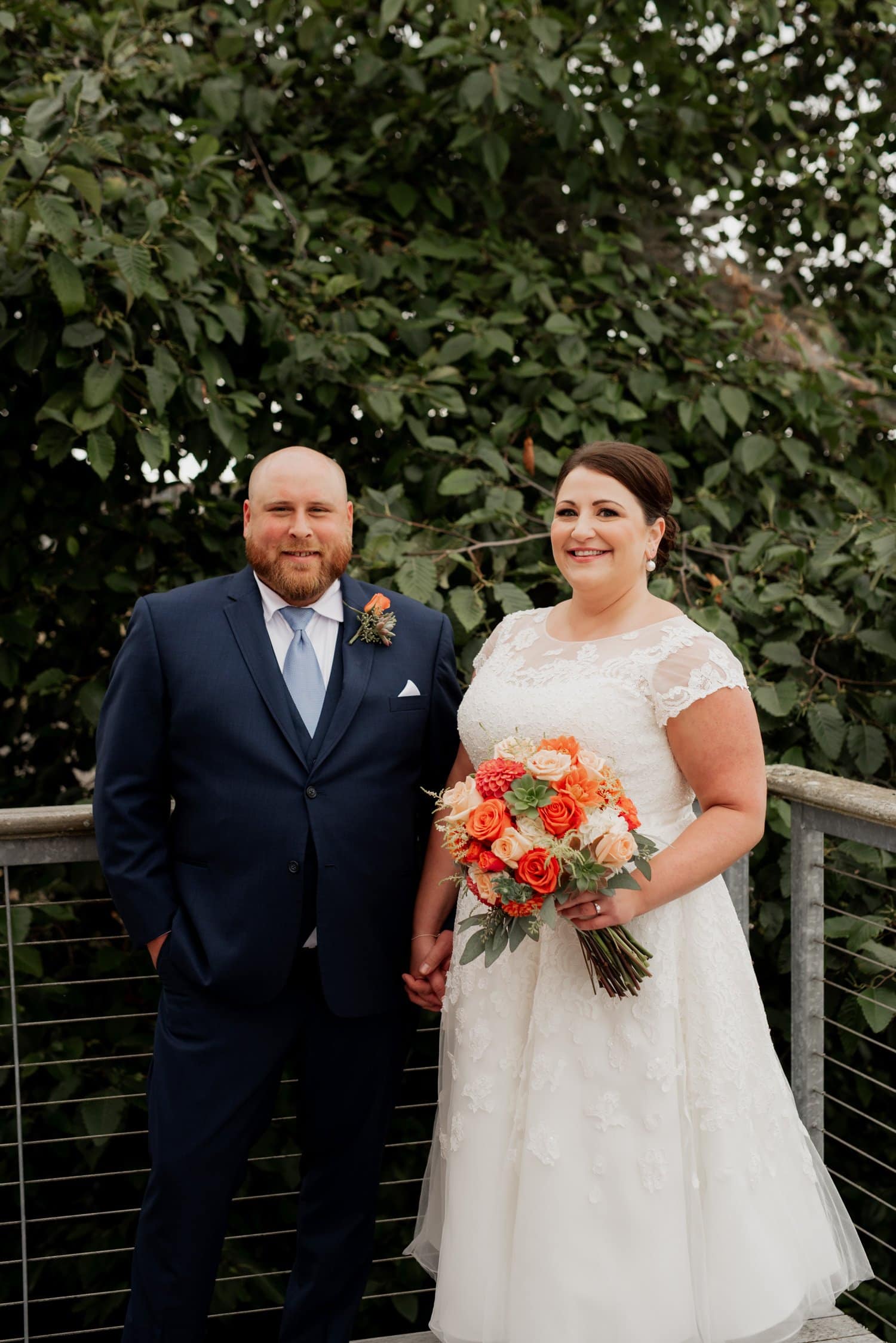 A couple in wedding attire holds hands, with the bride holding an orange bouquet, surrounded by lush greenery at The Loft at the Red Building.