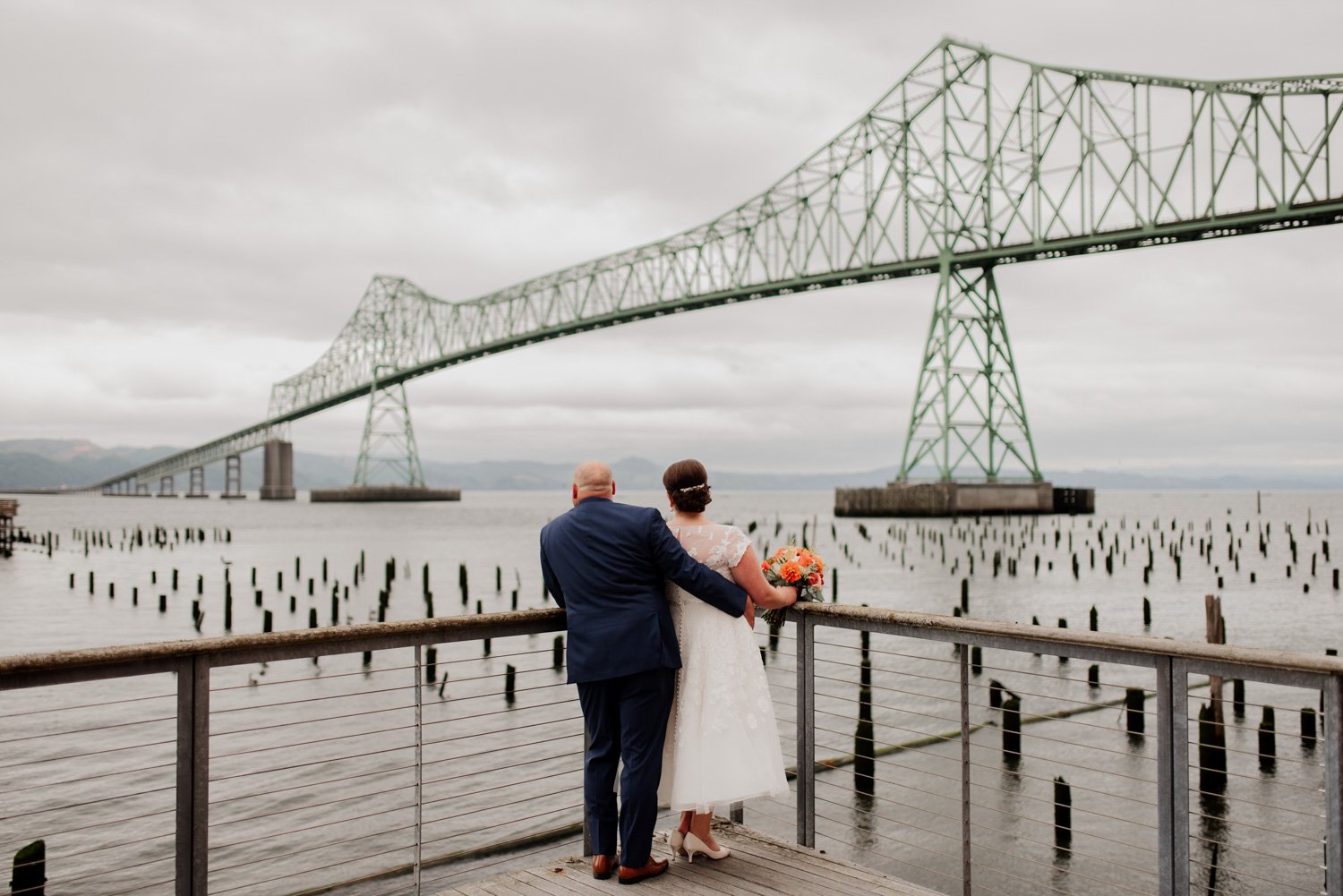 A couple in wedding attire embraces on a pier, with a large bridge visible in the background.