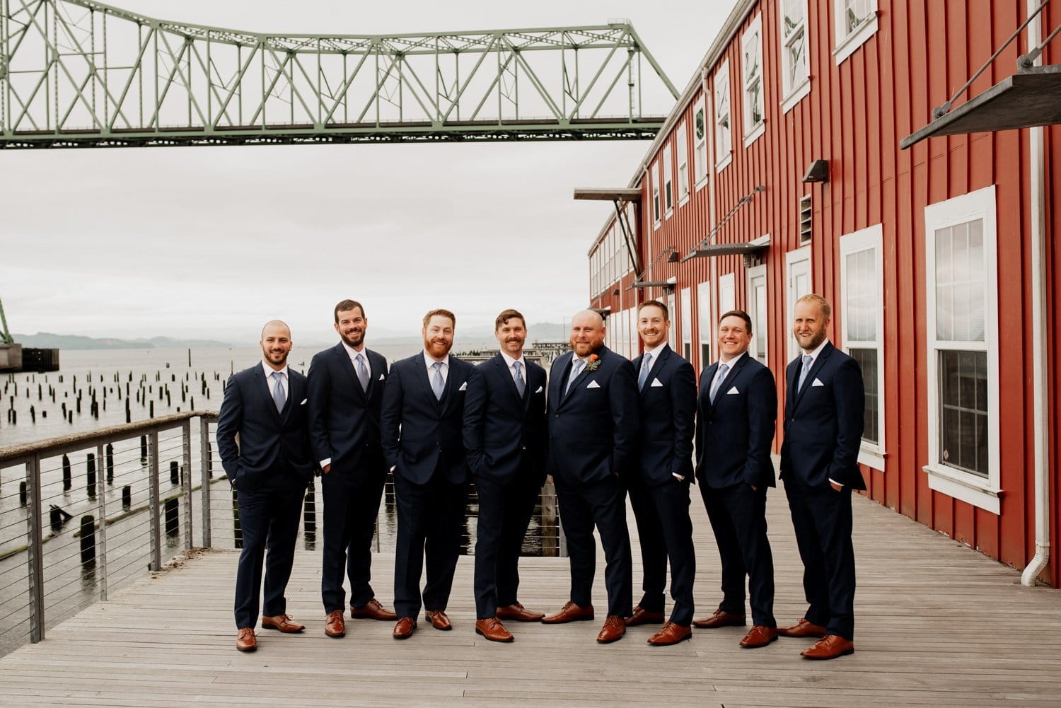 Eight men in suits stand on a wooden deck next to a red building, with a bridge and body of water visible in the background.