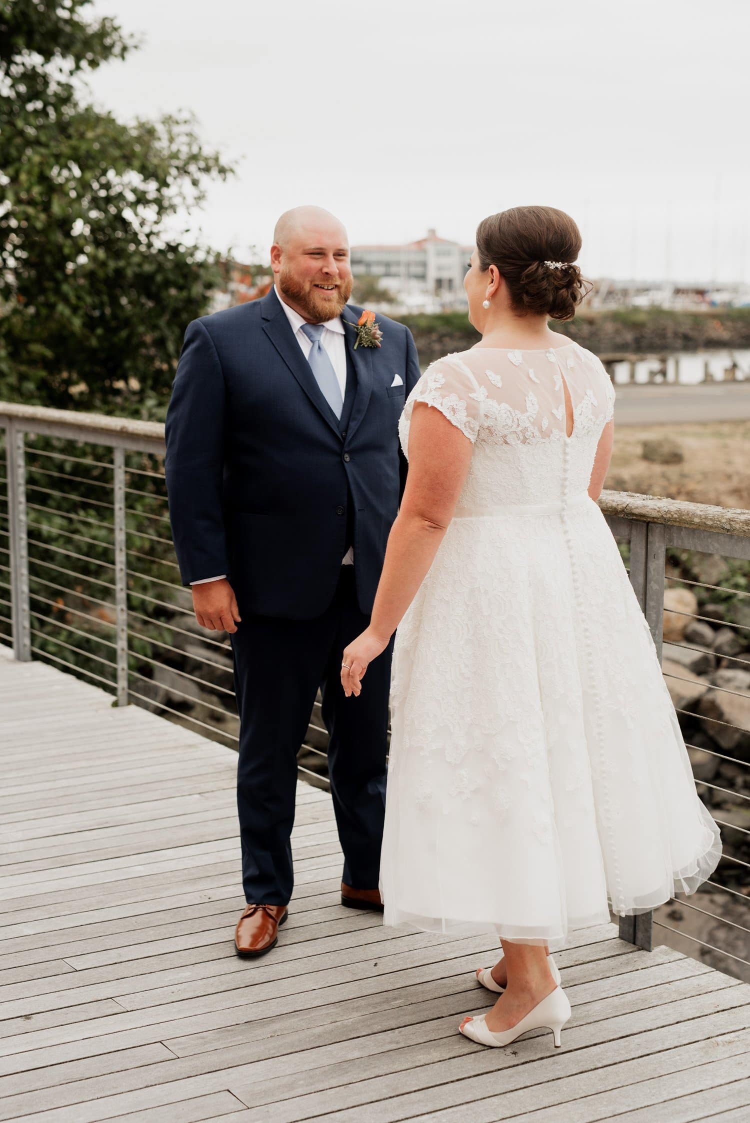 A couple smiles at each other on a boardwalk while capturing wedding photos. The bride is in a white dress, and the groom wears a dark suit, celebrating their special day at The Loft at the Red Building.