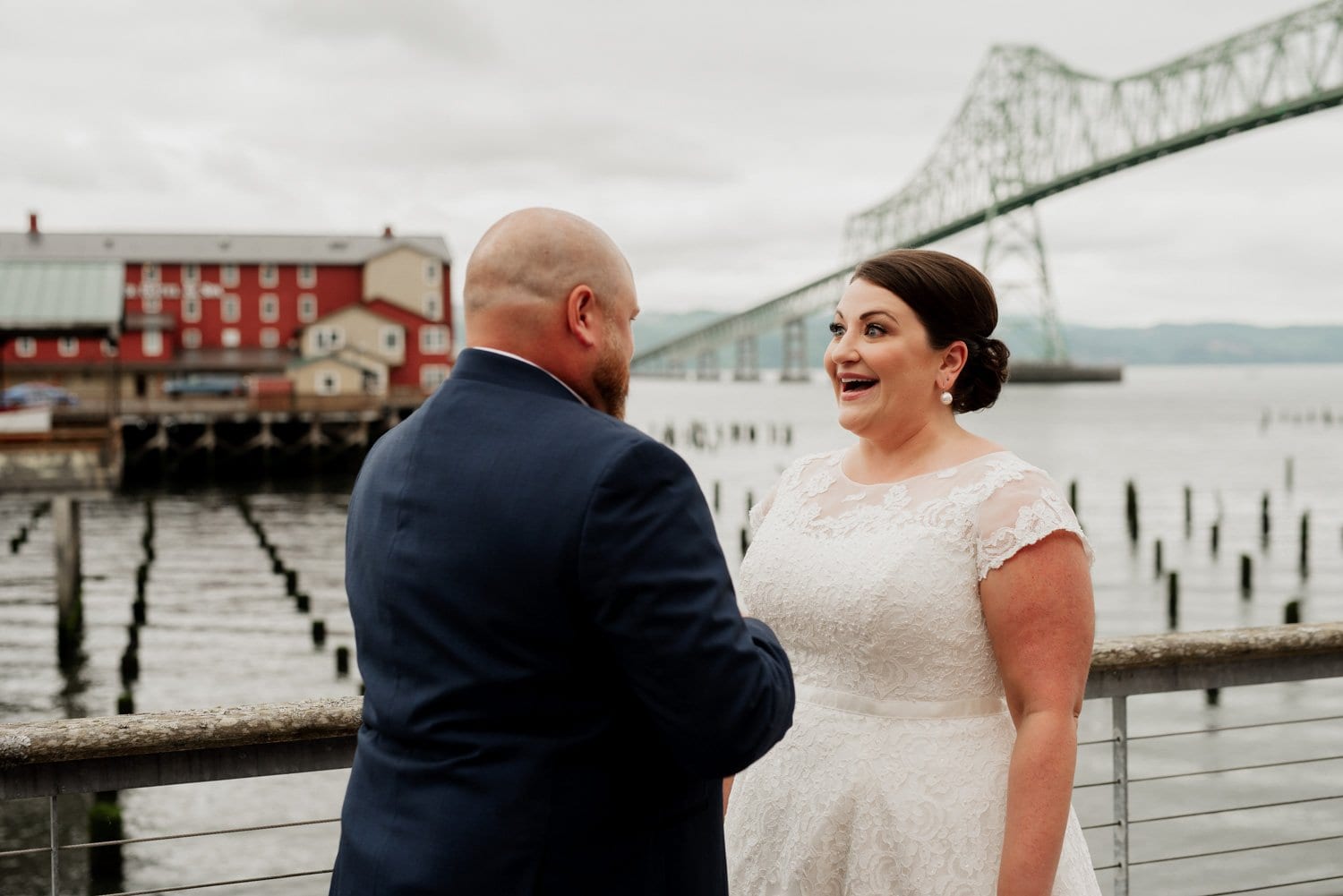 A couple in wedding attire shares a joyful moment by the waterfront with a bridge visible in the background, during their celebration at The Loft at the Red Building.