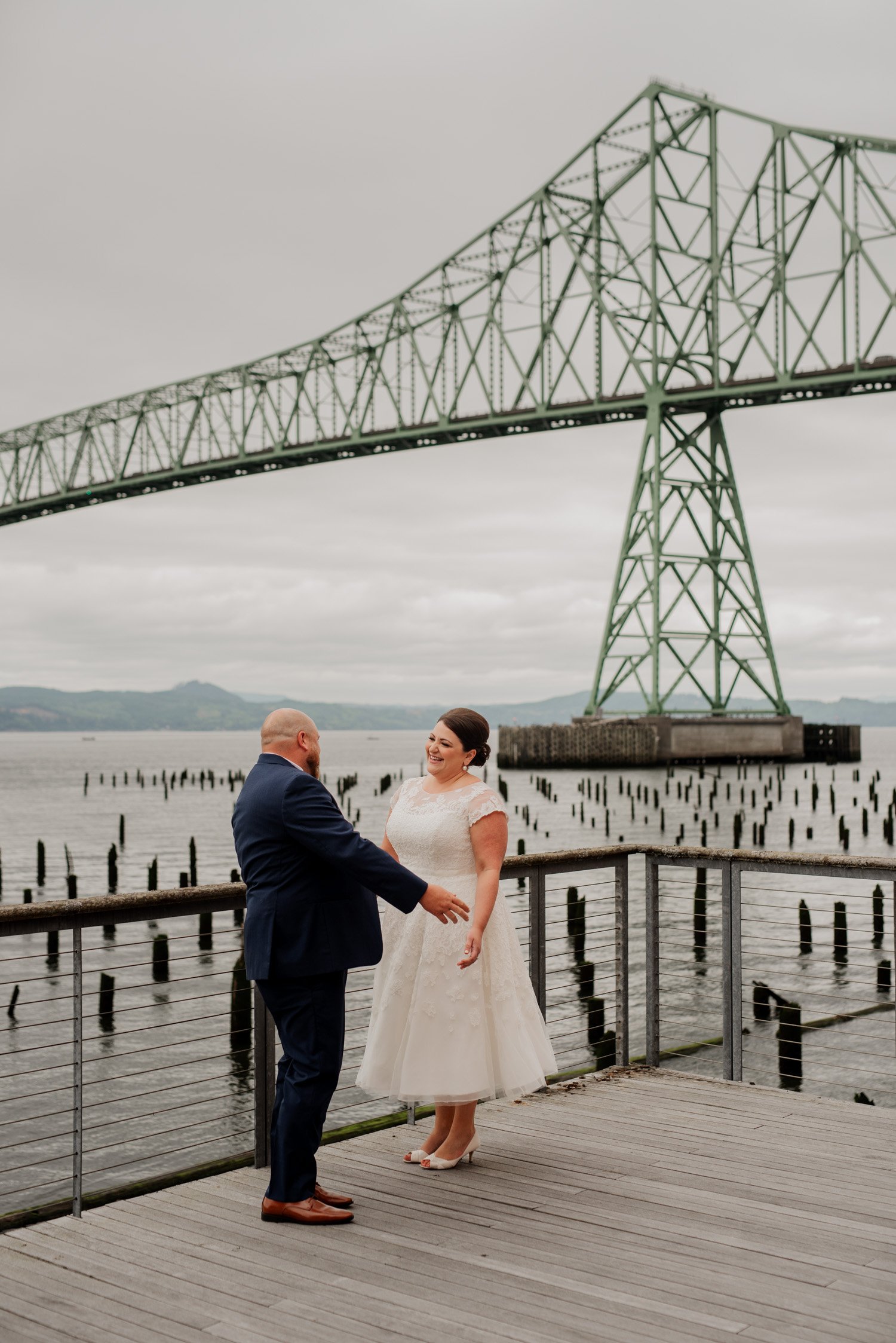 The bride and groom exchange joyful smiles on a dock while capturing beautiful wedding photos with a large bridge in the background.