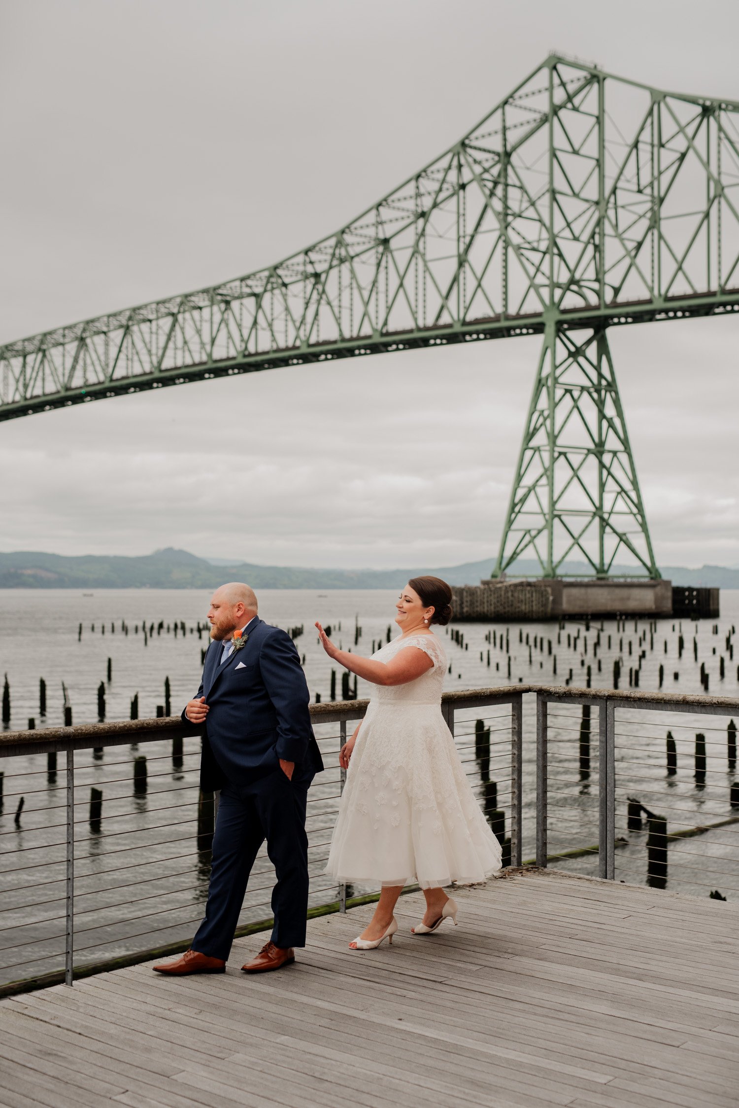 A bride playfully taps the grooms shoulder while standing on a boardwalk under a towering bridge. The couple is dressed in wedding attire, with the scene capturing a joyful moment from their special day at The Loft at the Red Building.