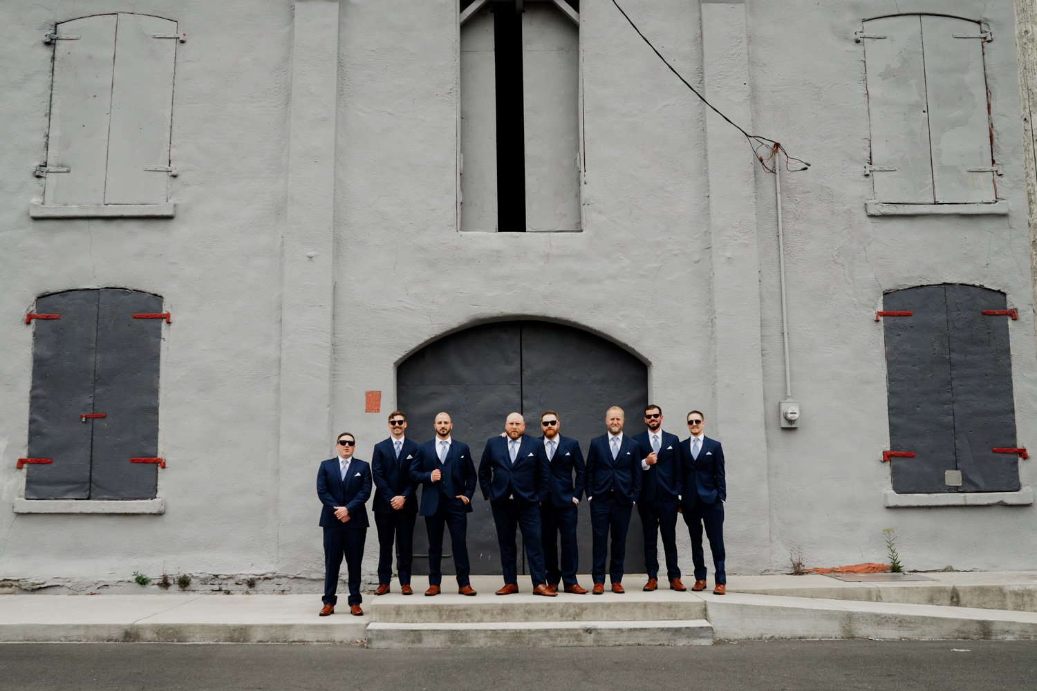 Eight men in navy suits stand in a row, posing for wedding photos against a large grey building in Astoria, Oregon.