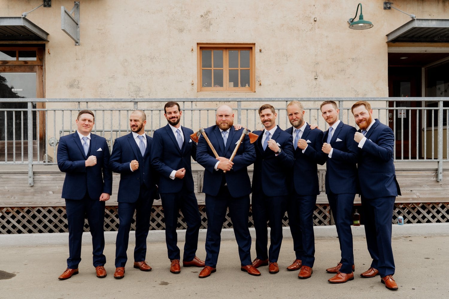 Eight men in navy suits and brown shoes stand confidently outside a beige building, showing camaraderie as they pose for wedding photos.