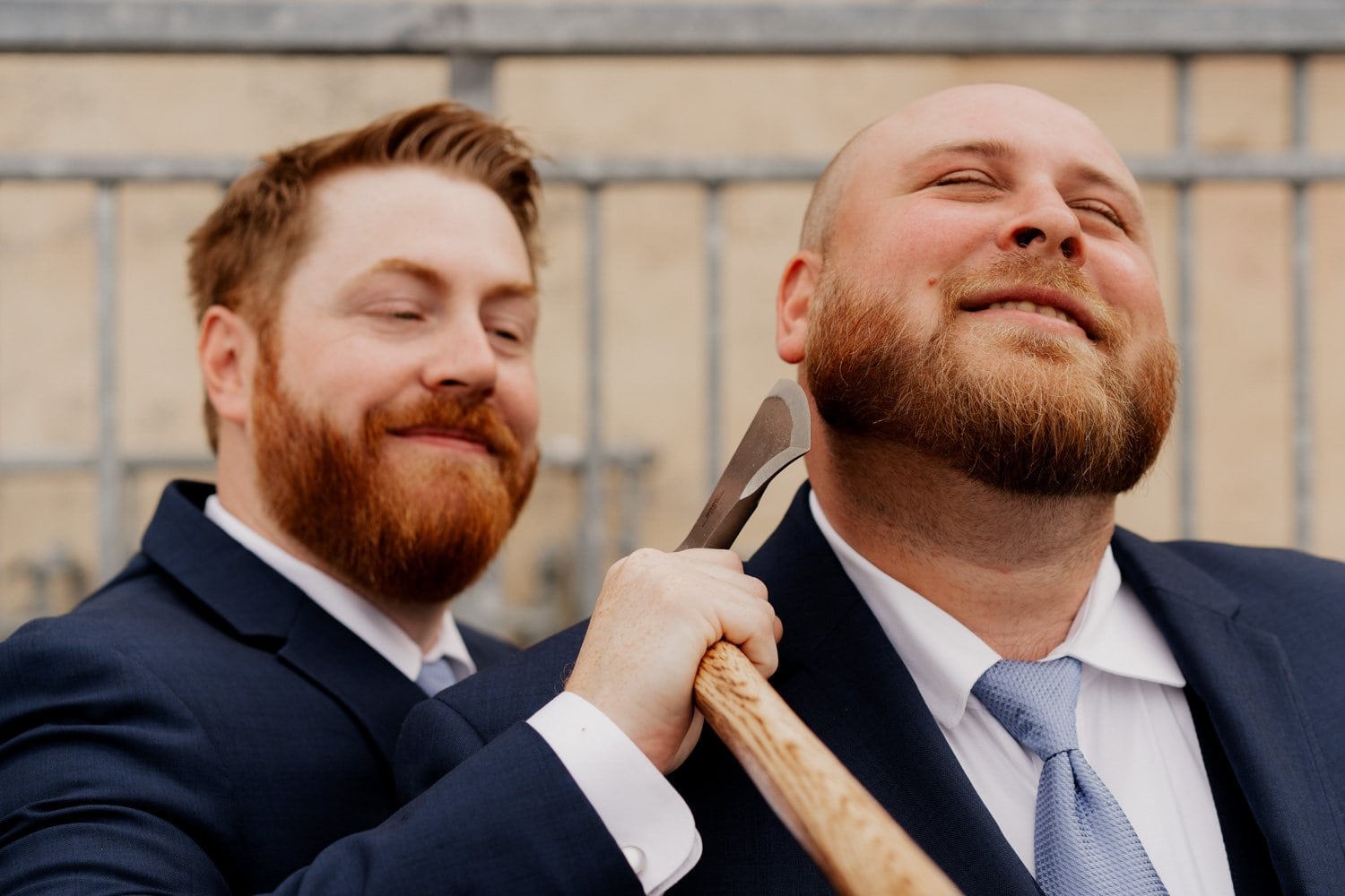 Two bearded men in suits smiling, with one pretending to shave the others beard using a small axe while getting ready at an Airbnb before a wedding. They are standing in a room decorated for the occasion.