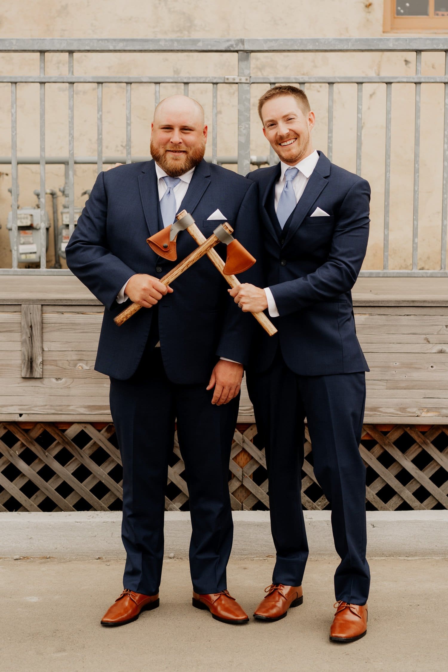 Two men in suits stand smiling, holding crossed axes in front of a wooden structure. This lively moment captures their excitement as they prepare for wedding photos.