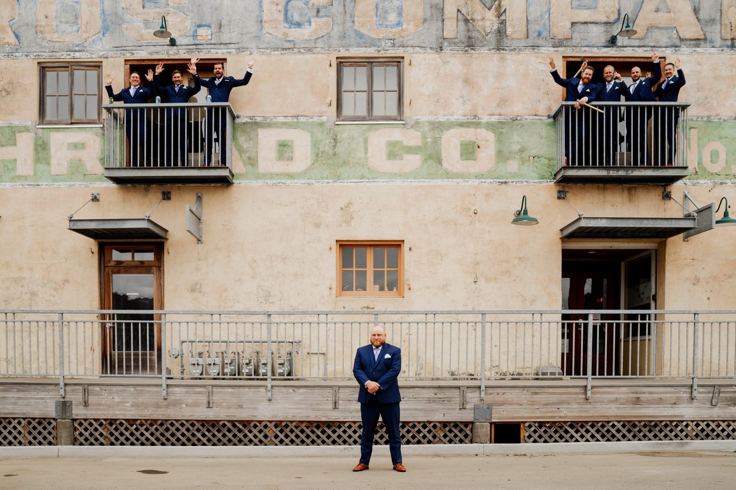 A man in a suit stands by a building, posing for wedding photos, while people in suits wave from the balconies above.