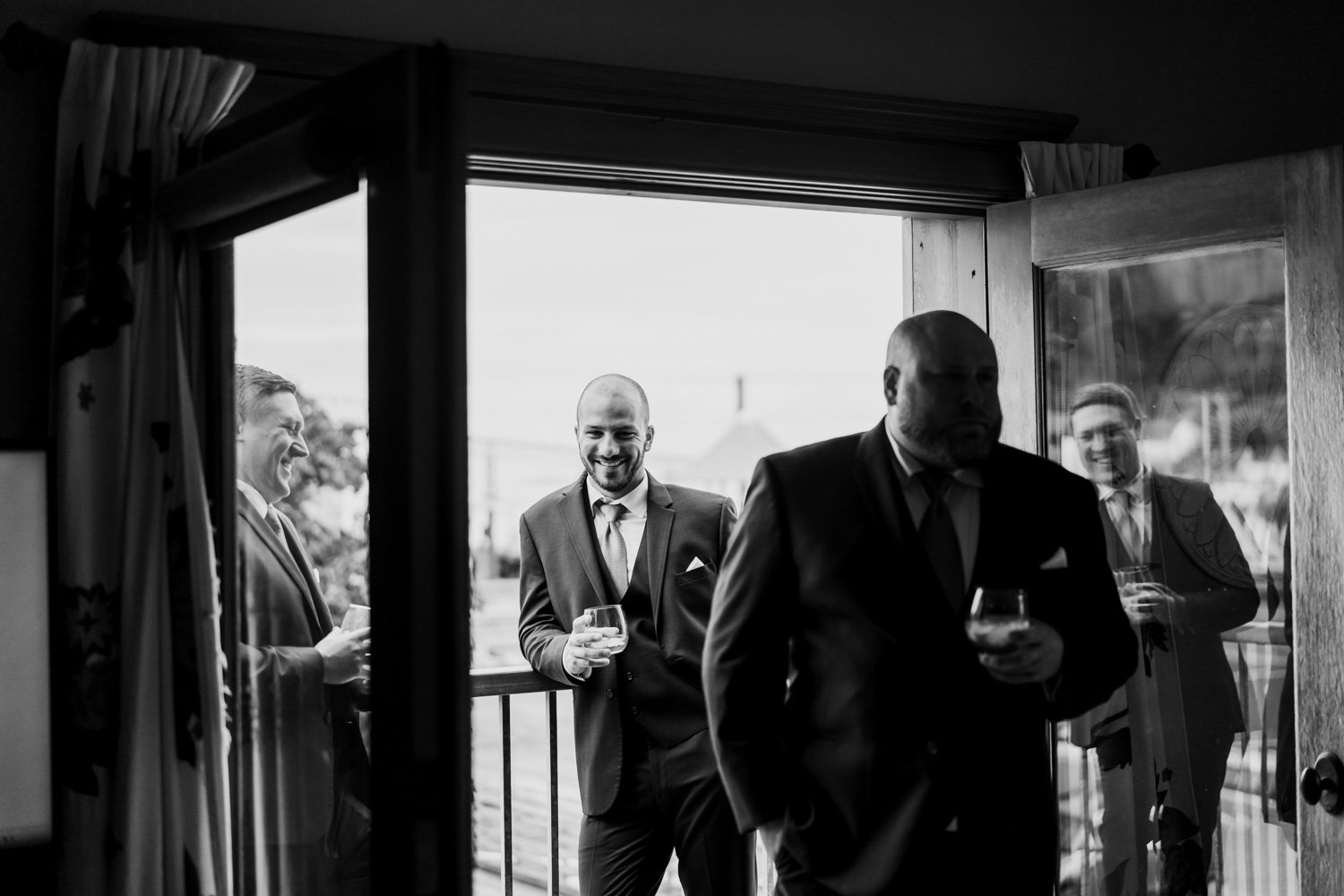 Four men in suits, two indoors and two on a balcony, smiling and holding drinks in preparation for a wedding. The photograph is black and white, capturing the men at an Airbnb before the event at The Loft at the Red Building.