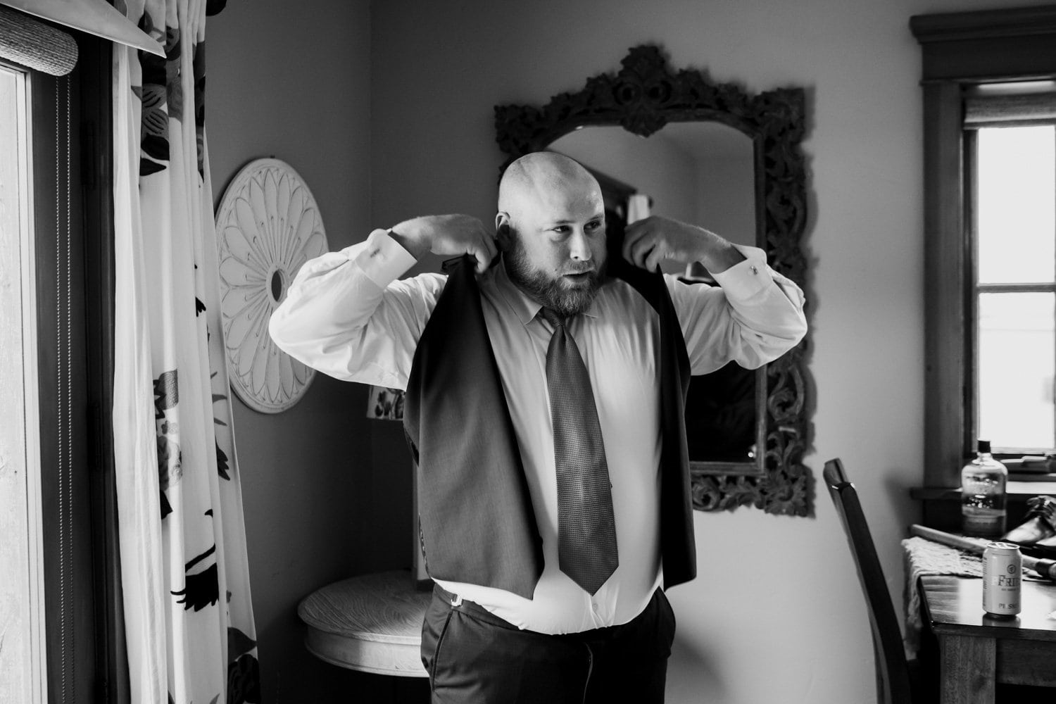 A man adjusts his vest in front of a mirror, with a curtain visible in the background. He is preparing for a wedding at The Loft at the Red Building while staying at an Airbnb.