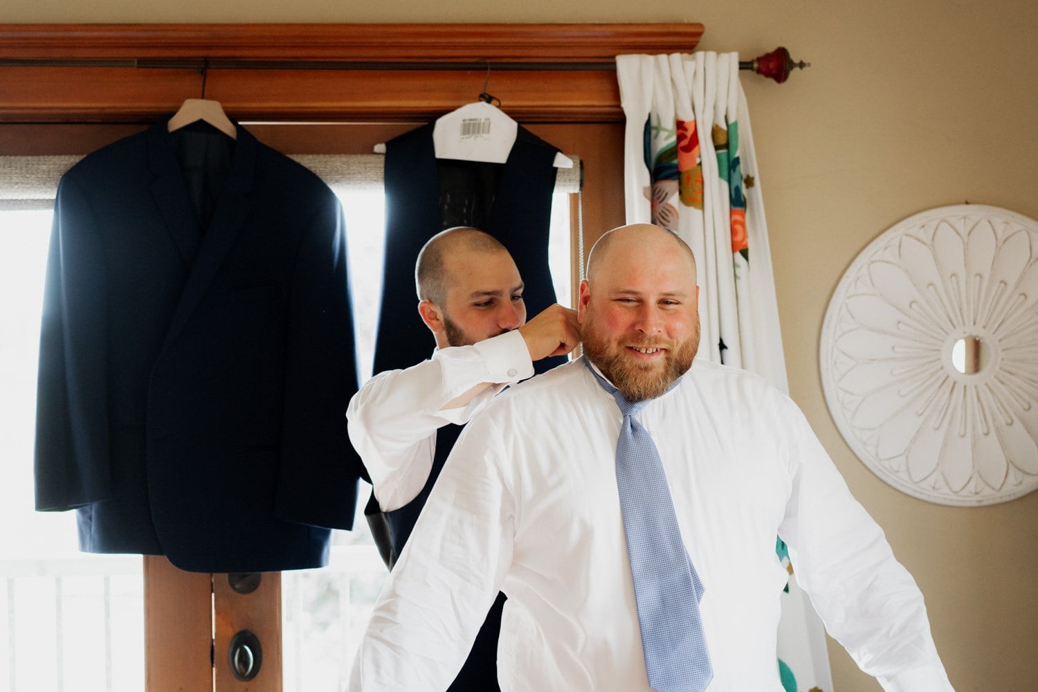 Two people in formal attire are preparing in an Airbnb room. One person is adjusting the other’s tie while a suit jacket hangs on a nearby chair. The setting has warm lighting, and photography equipment is visible in the background, indicating preparations for a wedding at The Loft at the Red Building.