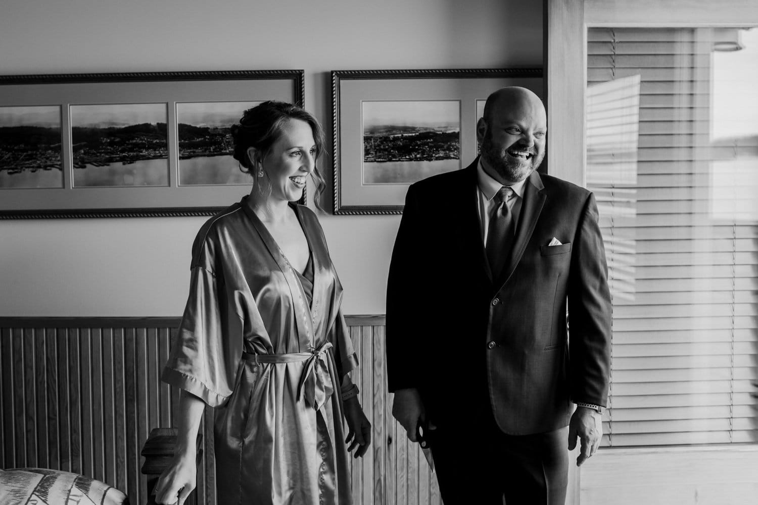 A smiling woman in a robe and a man in a suit stand indoors near framed pictures, preparing for their wedding photo session at The Loft at the Red Building.