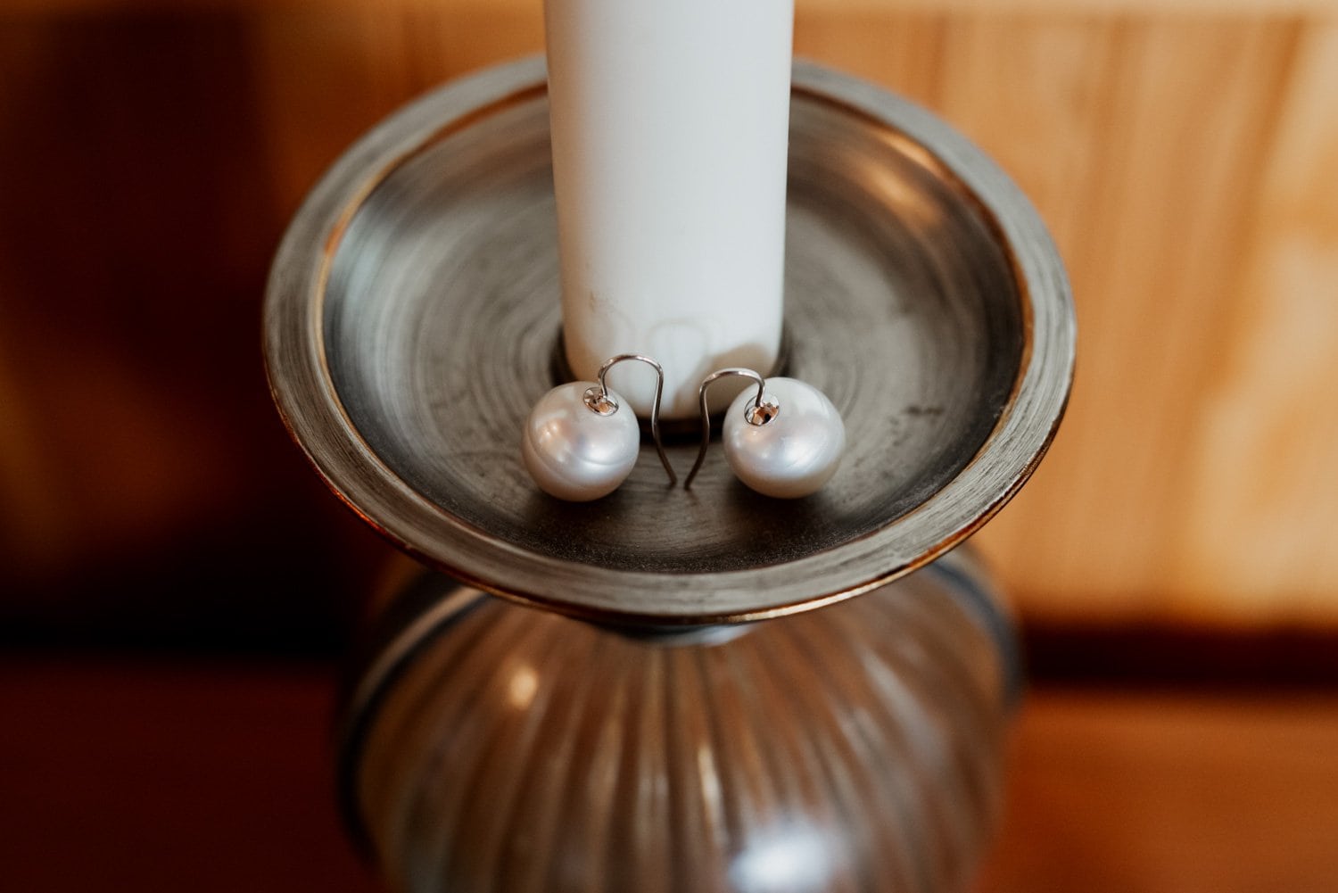 A pair of pearl earrings hanging from a silver candlestick next to a tall white candle, symbolizing preparation for a wedding.