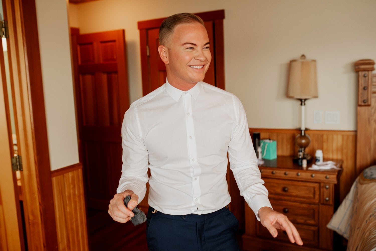 A smiling person in a white shirt holds an object in their hand while standing in a warmly lit room with wooden furniture, preparing for a wedding at The Loft at the Red Building.