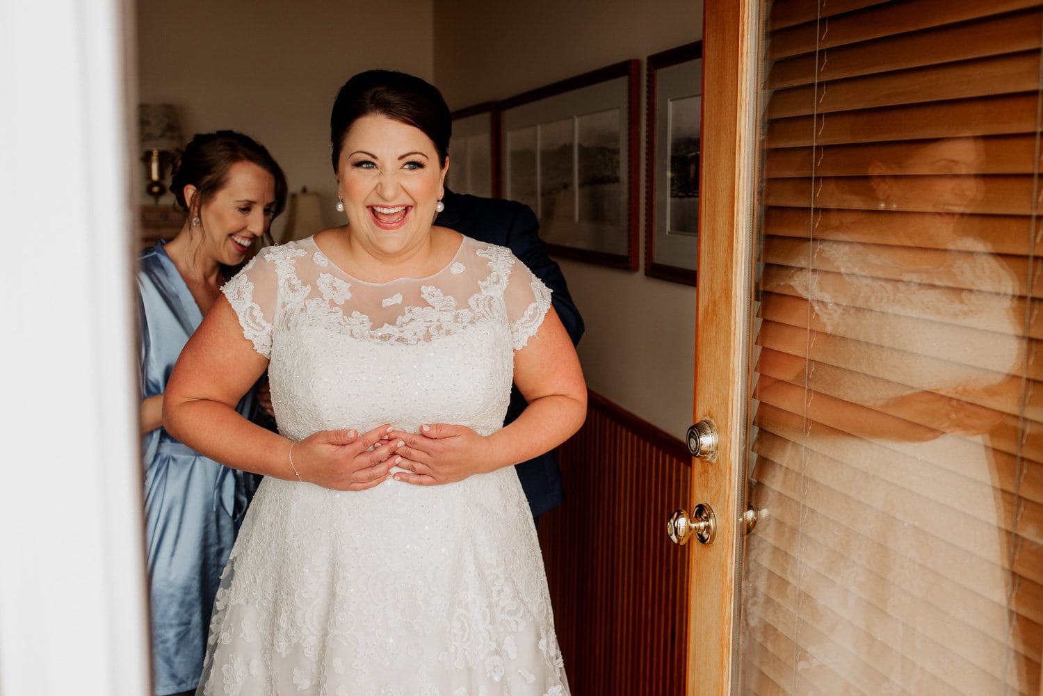 Bride smiling in a white lace dress as she prepares at an Airbnb, assisted by a woman in a blue robe.