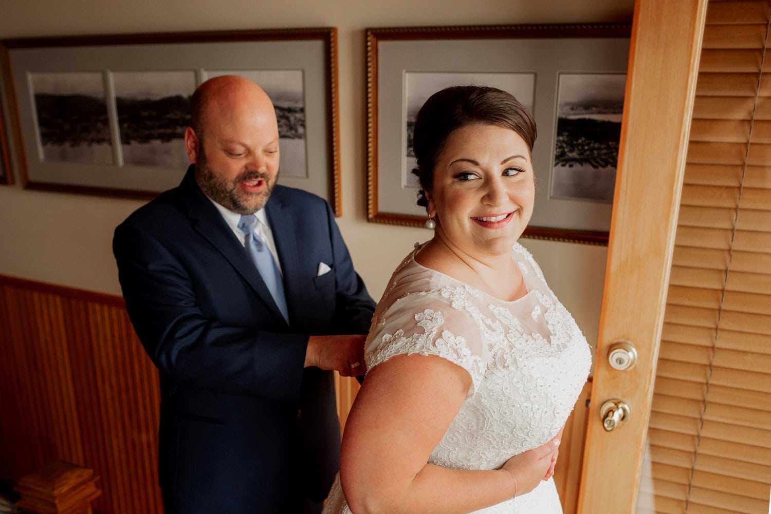 A smiling bride in a white dress stands by a door with framed wedding photos on the wall, assisted by a man in a suit, as they prepare for the wedding ceremony at The Loft at the Red Building.