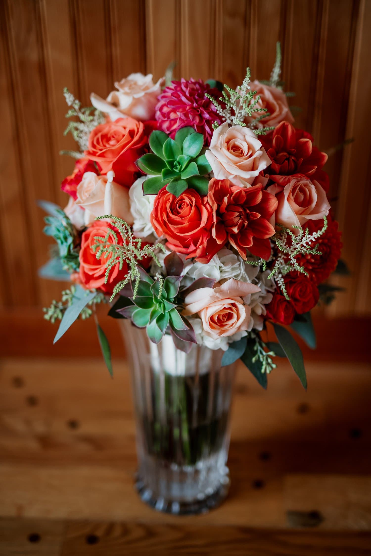 A vibrant floral arrangement consisting of red and pink roses, dahlias, and greenery is displayed in a glass vase on wooden floors in an Airbnb. The setting is prepared for wedding photos prior to the ceremony at The Loft at the Red Building.