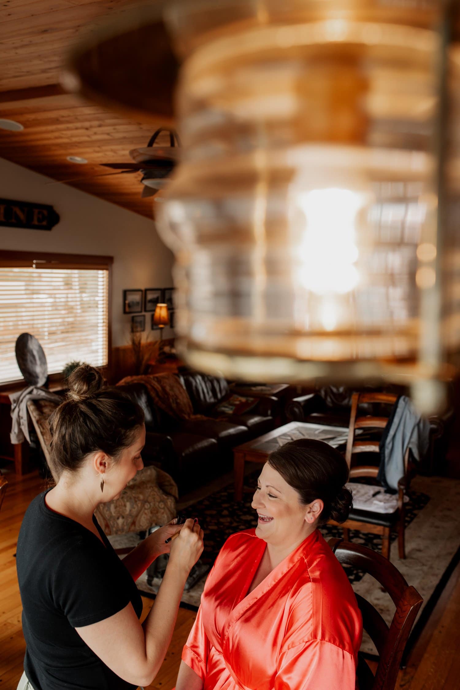 In a cozy Airbnb with wooden walls and a hanging light overhead, a woman in a red robe is having her makeup applied as she prepares for wedding photos at The Loft at the Red building.
