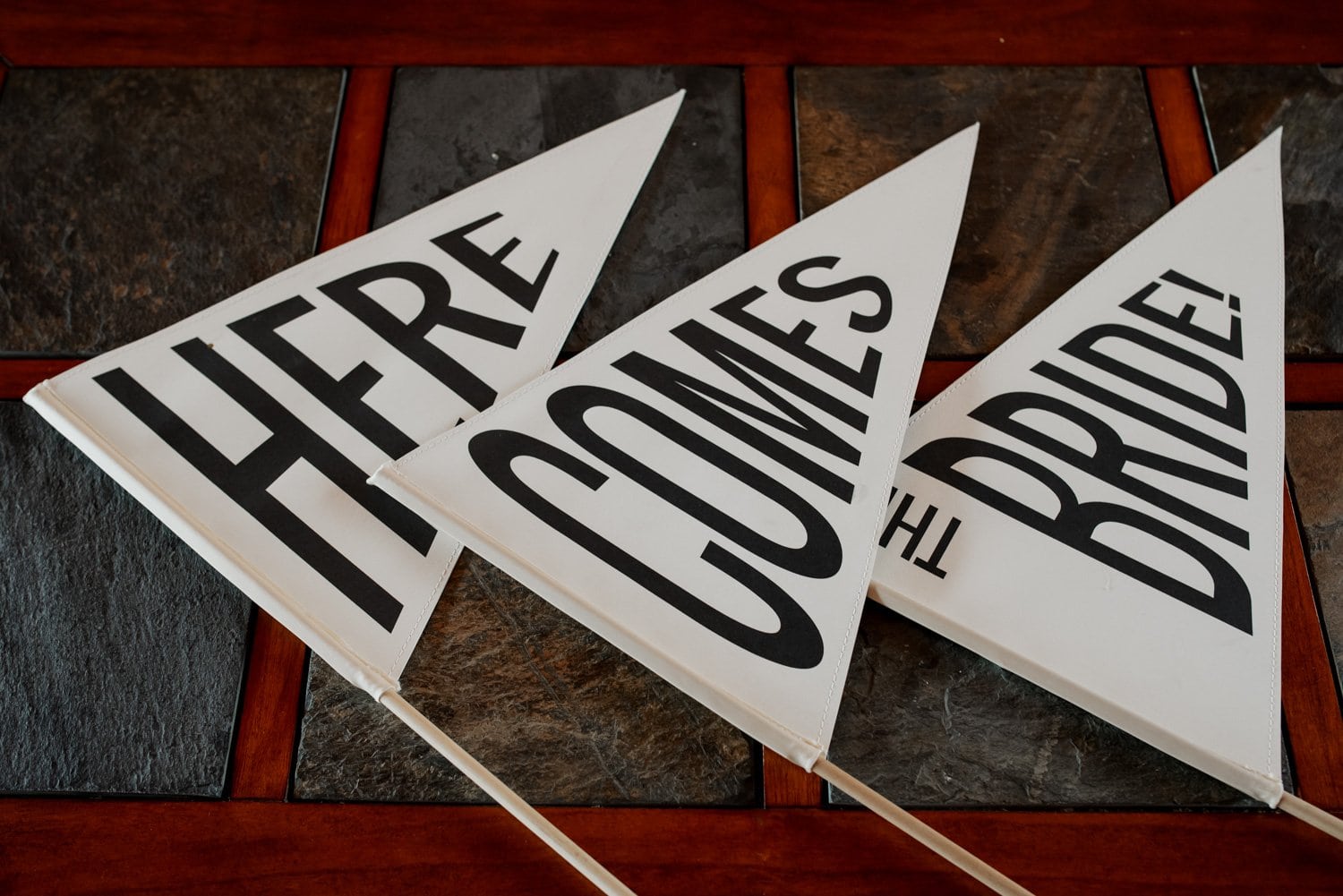 Three triangular flags on a tiled surface, featuring bold black letters that read Here Comes the Bride!.
