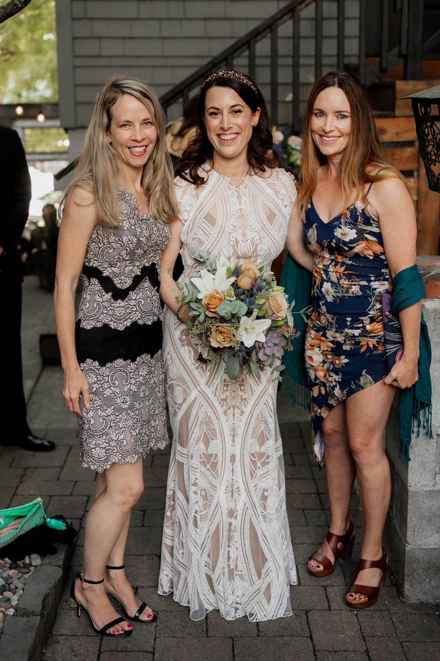 Three women standing outdoors at the wedding venue Opal 28, one in a white dress holding a bouquet and the other two in patterned dresses, all smiling.