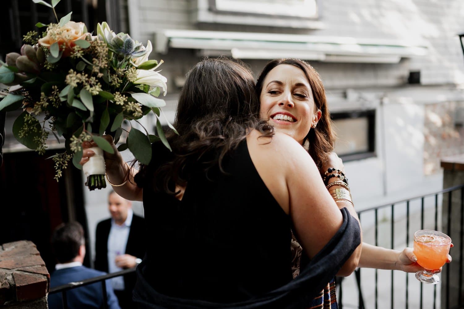 Two women share a warm embrace; one holds a bouquet, the other has a drink. Guests are chatting in the background at Opal 28 during a wedding.