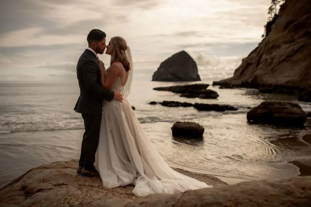 A bride and groom touch foreheads during their Cape Kiwanda elopement, standing on a rock overlooking the ocean with a large sea stack in the distance.